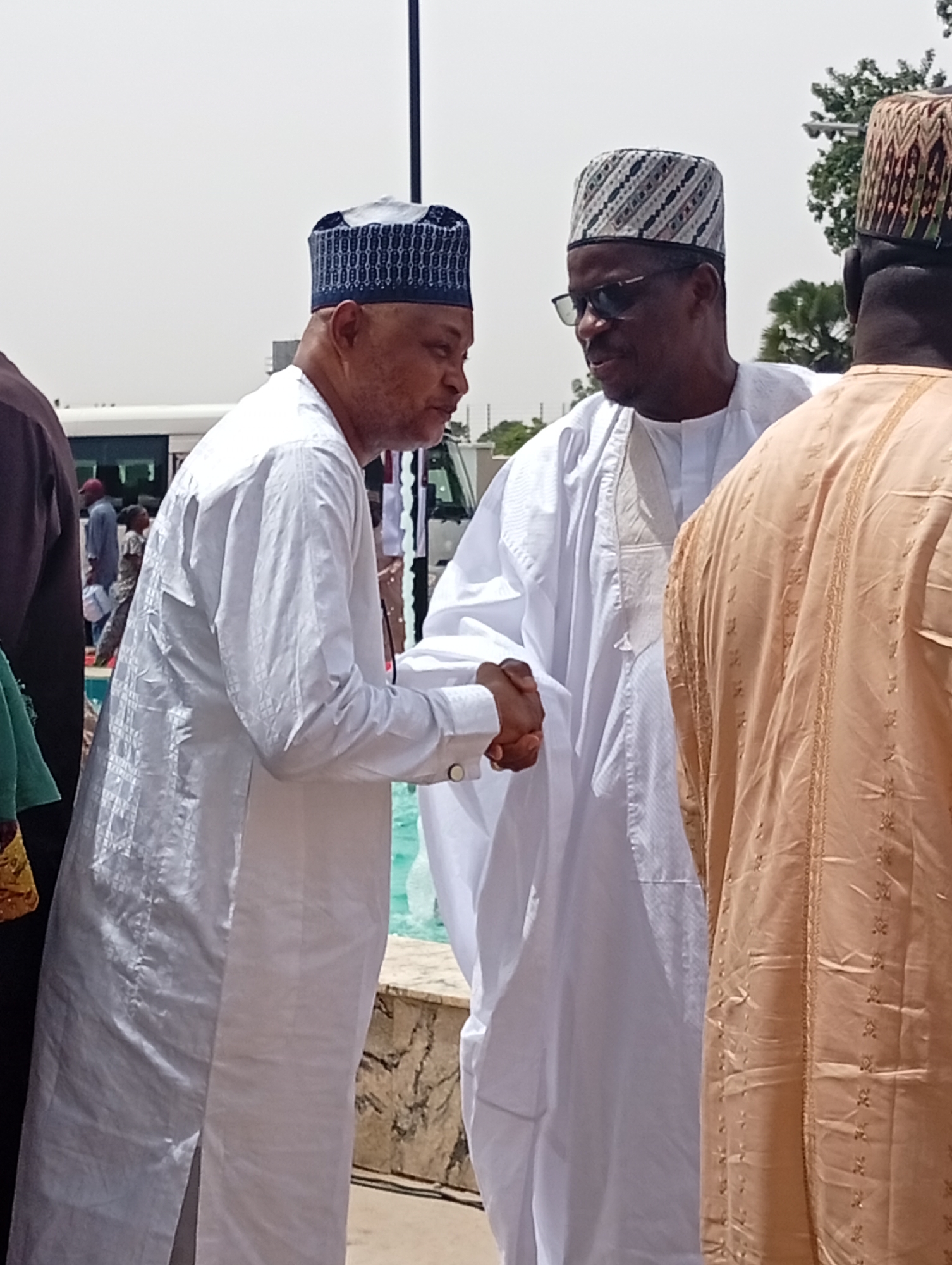 This bright outdoor image, set in Unguwan Sanusi, Nigeria, features two men in traditional attire shaking hands in the foreground, suggesting a greeting or formal interaction. The man on the left wears a white, long-sleeved tunic with a subtle checkered pattern and a dark blue and white embroidered cap. He faces the man on the right, whose hand he holds.
The man on the right is also dressed in a white, long-sleeved tunic and a patterned cap featuring black, white, and possibly brown geometric designs. He wears dark sunglasses and is looking towards the man on the left. To the far right, the back of a third man is visible, wearing a light brown or tan traditional garment with gold embroidery and a patterned cap similar to the man in white.
The scene is brightly lit by the sun, indicating midday or early afternoon, with clear skies. In the mid-ground, behind the handshake, there is a body of water, likely a fountain or a pool, with a decorative stone edge. A light-colored bus or van is partially visible in the left background, while distant trees and foliage, along with a tall dark pole, are seen on the right. The overall impression is a cordial gathering or event in an open, sunny outdoor space. No text is visible in the image.