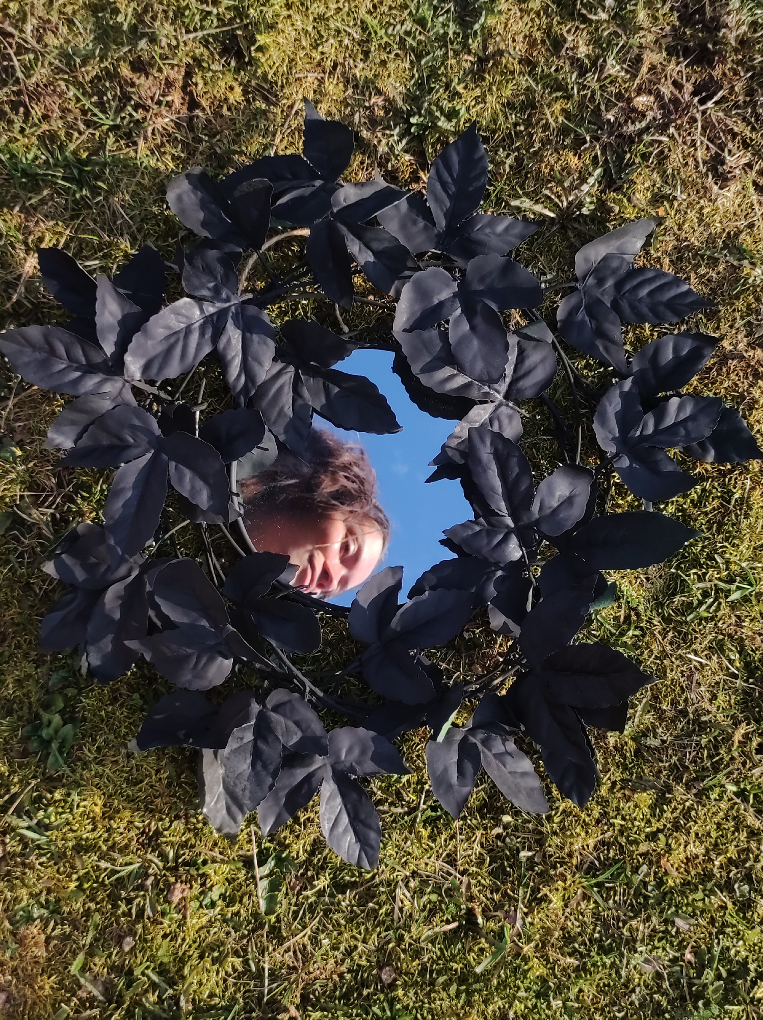 A top-down shot depicts a round mirror placed on a grassy and mossy surface. The mirror's frame is adorned with black leaves forming a wreath. Reflected in the mirror is the face of a person under a blue sky. The location cannot be confirmed based on the image.