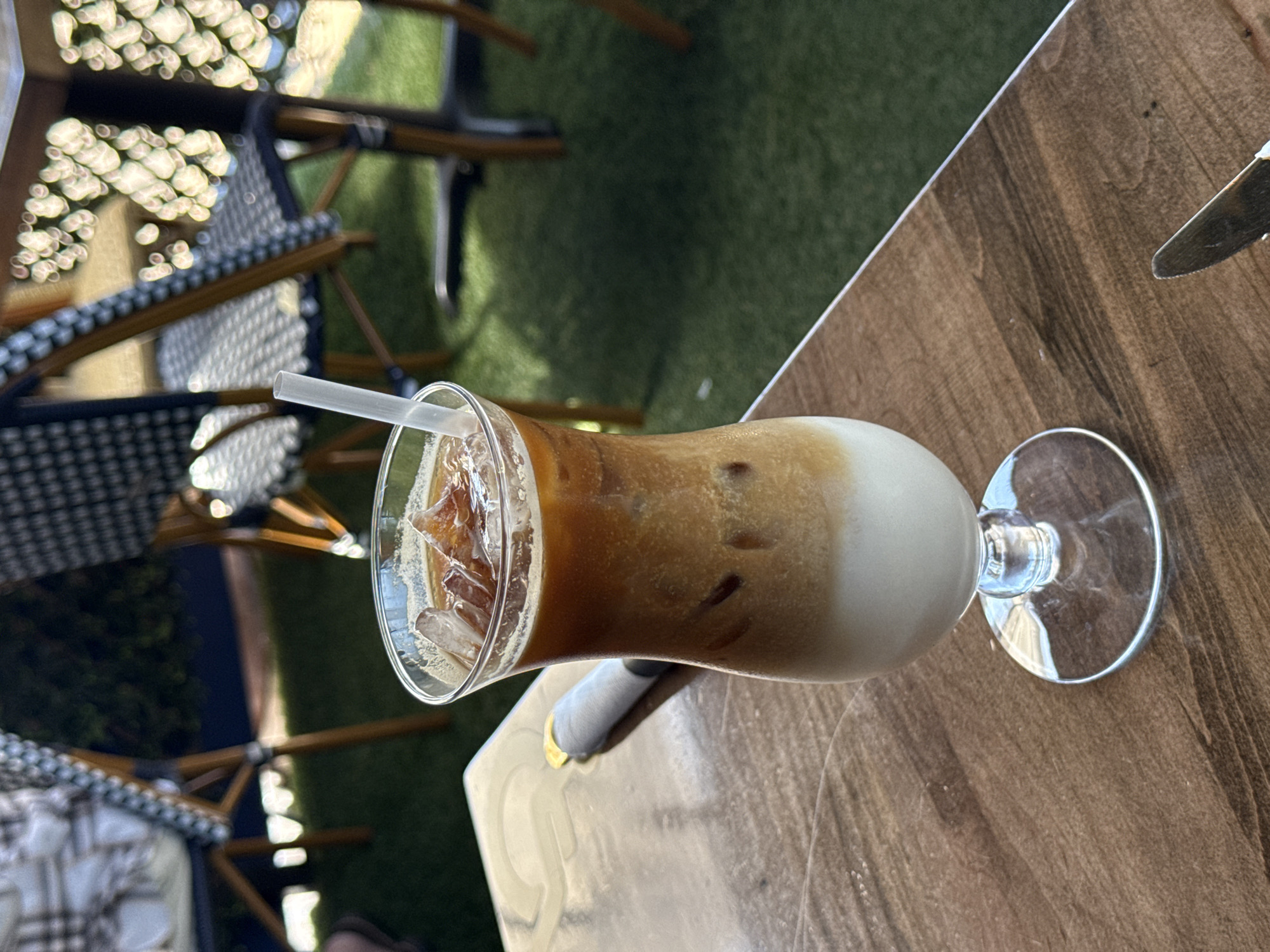 The image shows a cocktail glass filled with an iced coffee drink, positioned on a wooden table outdoors. A white straw is in the drink, which appears to be layered with ice, coffee, and milk or cream. A knife is visible on the table next to the glass. In the background, there are chairs and a green grassy area. The location is likely in Chula Vista, United States of America.