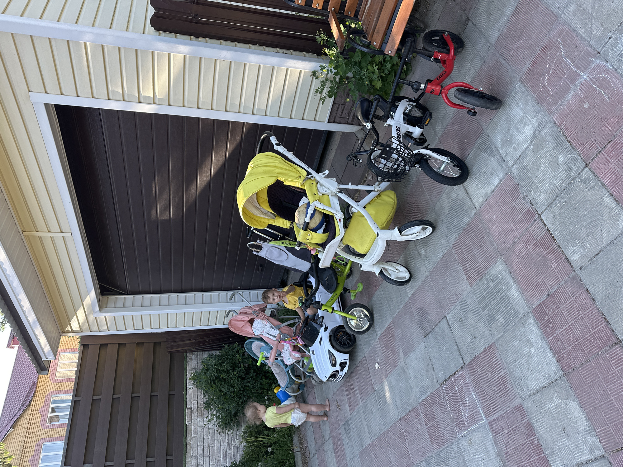 The image depicts a scene in Nizhny Novgorod, Russia, on a paved area composed of red and gray bricks, likely a private residence exterior. There are four children's vehicles parked near the exterior wall of a house: a yellow stroller, a pink stroller, a white toy car, and a white and red bicycle. Two small children, both wearing light-colored clothing, are present; one is standing near the bottom edge of the image, and the other is sitting on the white toy car. There is also a possible third child in the Yellow stroller. A bench sits atop the vehicles. The exterior wall of the house is paneled with two colors of siding, tan and dark brown. Shrubbery and foliage are visible growing along the base of the fence and near the bench. A portion of another house with brick siding can be seen in the upper left corner.
