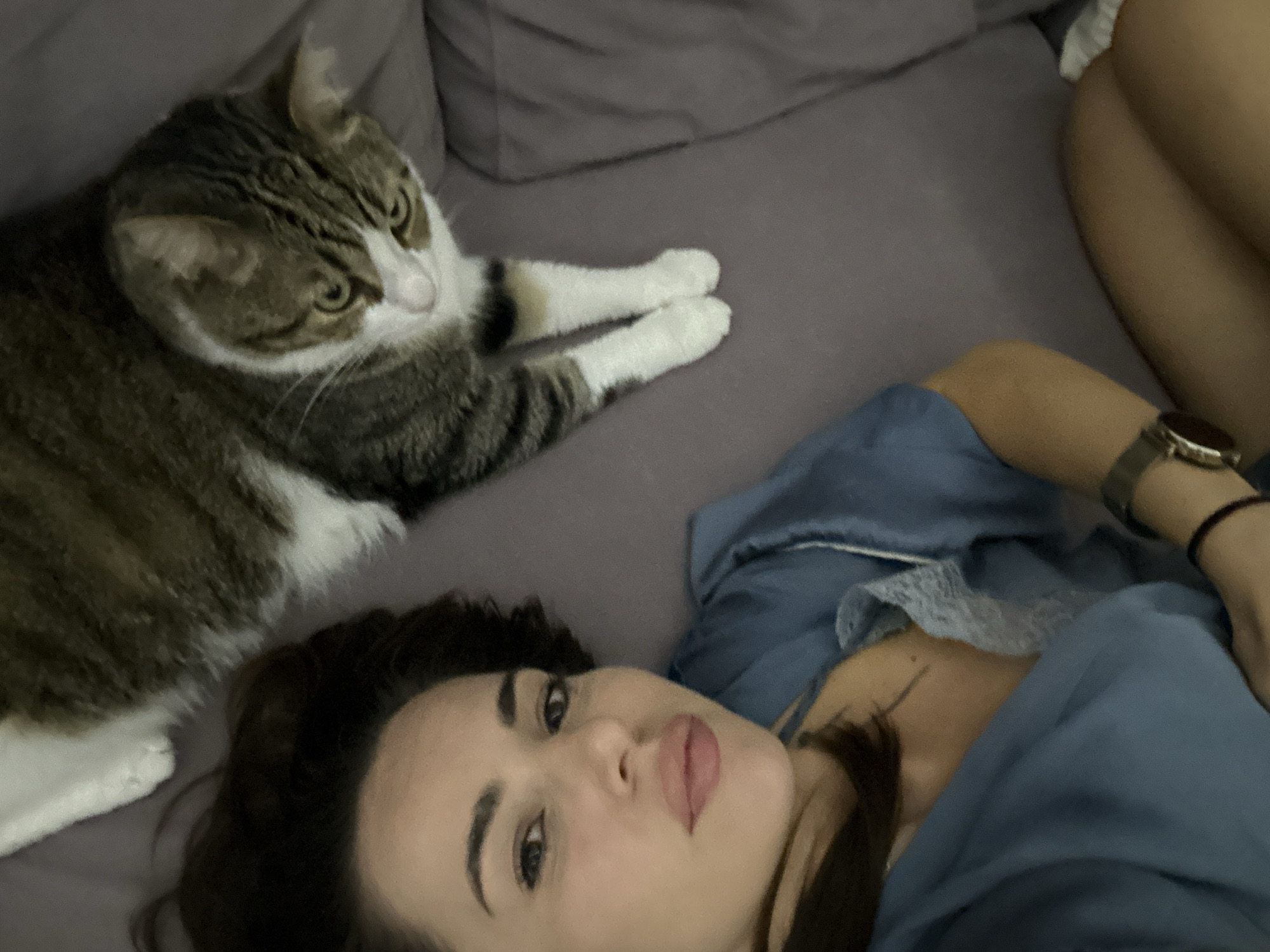 An indoor, top-down shot features a person and a cat lying on a grey, soft surface, likely a bed or sofa. In the foreground, a woman with dark hair and a neutral expression looks directly at the camera. She wears a blue top, possibly loungewear, with a hint of white lace trim. A watch with a metallic band is visible on her left wrist, and a dark bracelet is on her right. Her lips are slightly parted.
To the left of the woman, a domestic cat with tabby (brown and grey striped) and white fur is resting. The cat's head is turned to the right, looking away from the camera, with its eyes appearing focused elsewhere. It has prominent white markings on its face, chest, and paws. Its front paws are extended forward on the grey surface.
The lighting is soft and subdued, suggesting an indoor setting during evening or night. The background is a continuation of the grey bedding or upholstery. The overall scene conveys a sense of casual relaxation and companionship. There is no visible text within the image.