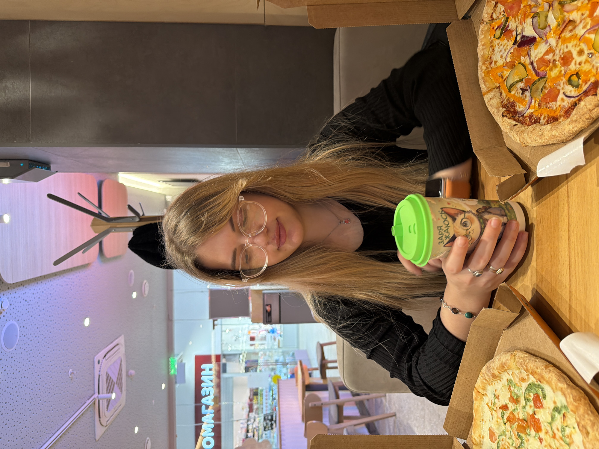A young woman with long, blonde hair and glasses sits at a wooden table in a brightly lit interior, likely a restaurant in Naberezhnye Chelny, Russia, indicated by the Cyrillic text on the store sign visible through the window. She is wearing a black, long-sleeved shirt. Two open pizza boxes frame her, one containing a pizza with a variety of toppings, including what appear to be onions, pickles, tomatoes and a yellow sauce, and the other containing a pizza with green toppings and tomatoes. In her hands, she holds a paper cup with a green lid and a cartoon illustration. She is wearing a bracelet with stones. The setting includes modern lighting fixtures and minimalist furniture.