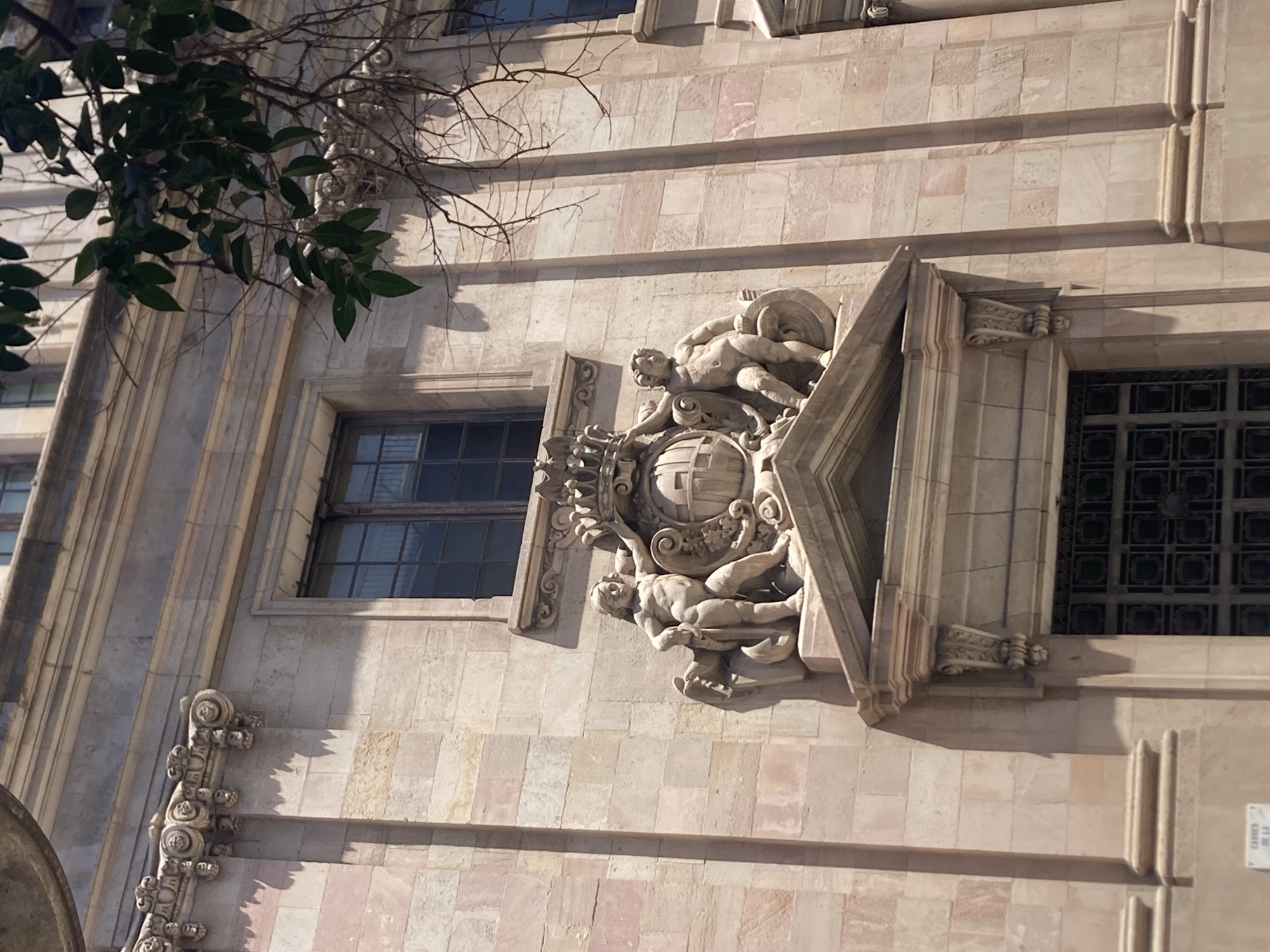 The image shows a close-up of the facade of a building in Barcelona, Spain. The architecture features large, light-colored stone blocks with vertical pilasters. A prominent decorative element is centered on the facade, depicting two cherubic figures flanking a globe with a crown on top. One cherub is on the left, holding a cloth, and the other is on the right, leaning on the globe. Above this sculpture, a rectangular window with dark panes is visible. The building is bathed in sunlight, casting shadows. A tree branch with green leaves hangs from the upper right corner of the frame, partially obscuring the building's upper section. The bottom of the image shows the top of a large, ornate door with a dark, lattice-like metal grate.