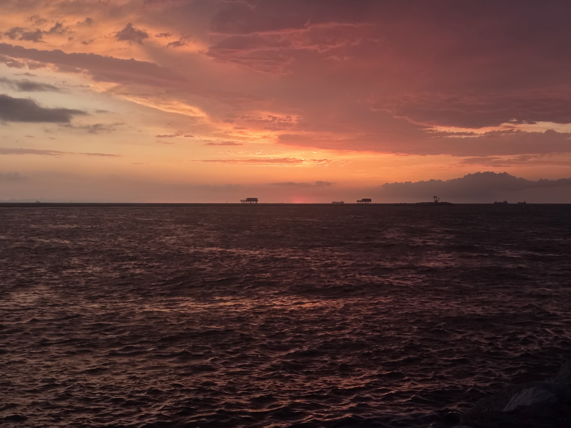 The image shows a sunset over the sea, with the sky painted in hues of orange, pink, and purple. The water is dark and choppy, reflecting the colors of the sky. In the distance, silhouetted against the horizon, are several structures that appear to be stilt houses or small piers. One structure on the left has a distinct roofline, while others are simpler in design. There are also faint outlines of ships or boats further out at sea. The overall impression is one of a peaceful and serene evening by the water.