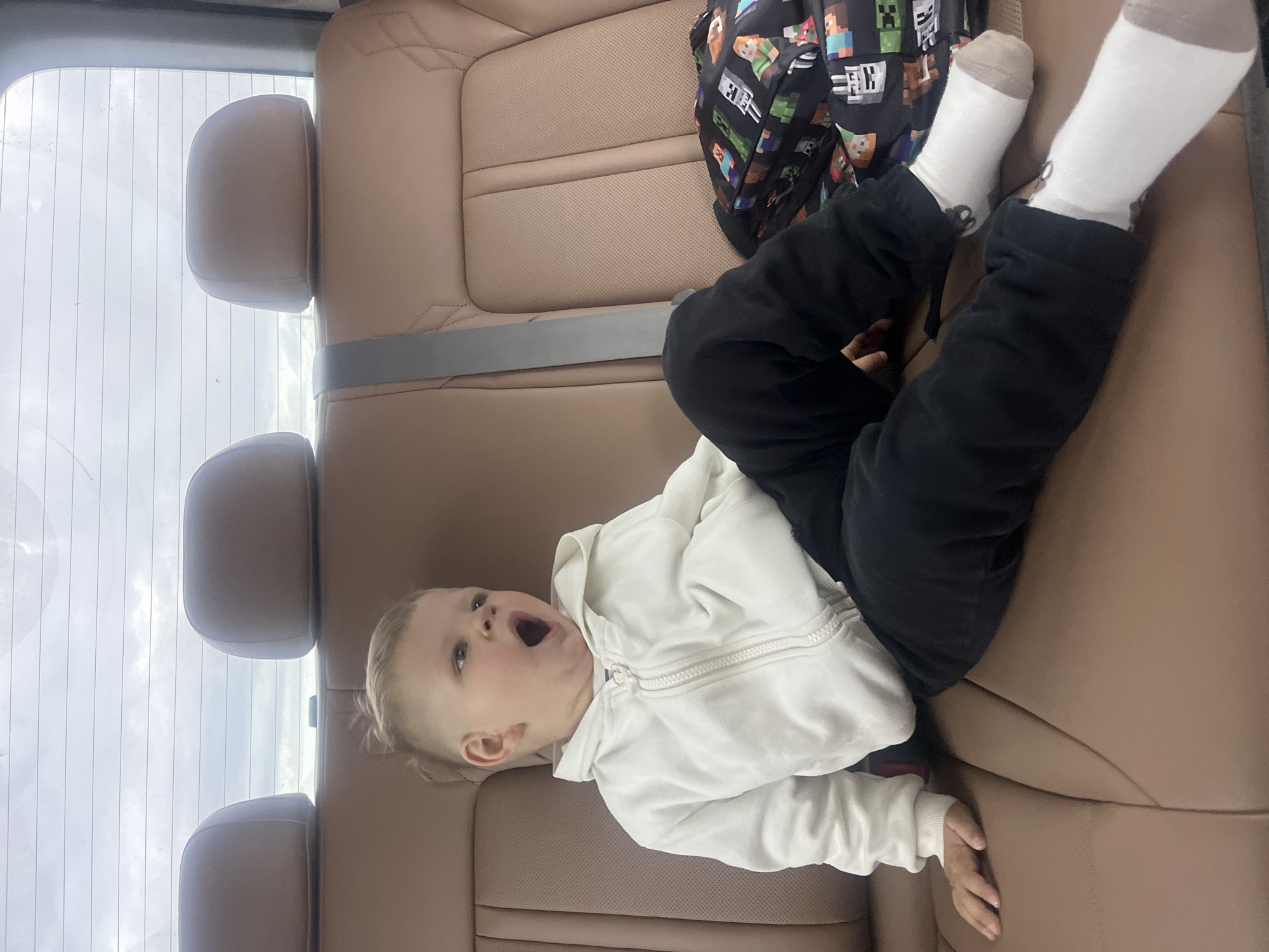 A young child with blonde hair is sitting in the back seat of a vehicle. The child is wearing a white zip-up hoodie and black pants. They are yawning widely, with their mouth open and eyes partially closed. A backpack with a Minecraft pattern is visible next to the child. The car's interior is upholstered in brown leather, and headrests are visible in the background, along with a portion of the rear window showing a cloudy sky. This scene is not located in Dybshche, Ukraine.