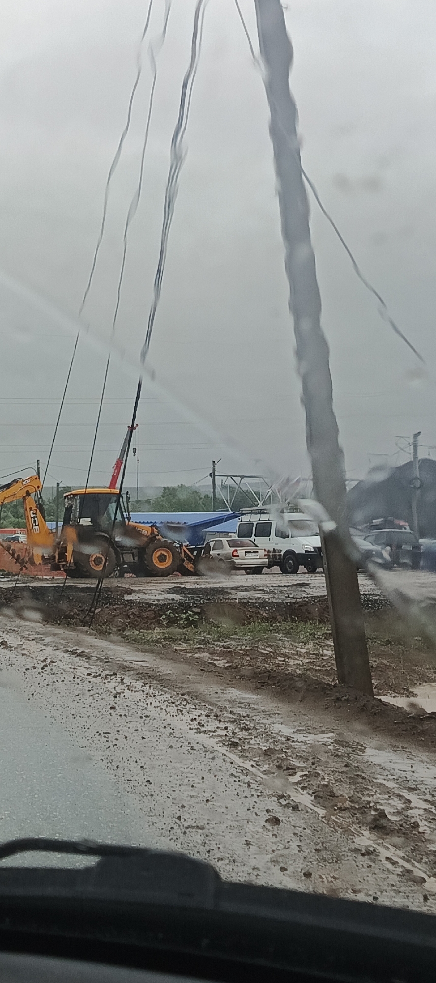 The image captures a construction site during what appears to be a rainy or overcast day, possibly in Kabanikha, Russia. The foreground is dominated by a wet, muddy road or ground, with water pooling and reflecting the dull sky. Several vertical lines, likely raindrops on a window or windshield, obscure parts of the scene.
The primary subjects are construction vehicles and other vehicles. A bright yellow JCB backhoe loader is prominently positioned on the left, engaged in some form of digging or earthmoving activity, indicated by the disturbed soil. Next to it, a smaller crane is visible, with its arm extended upwards. Further to the right, a white van and a light-colored sedan are parked or stopped, partially submerged in mud. In the background, more vehicles and construction equipment are present, along with some blue tarpaulin sheeting. Power lines and utility poles are also visible against the cloudy sky, suggesting an urban or semi-urban setting undergoing development or repair.
The activity taking place is clearly construction work, possibly involving road maintenance or building in challenging weather conditions. The mud and water suggest recent or ongoing precipitation. The time of day is not definitively clear but the overcast sky points towards daylight hours, likely in the morning or afternoon. The overall mood is somber due to the weather, but the presence of active machinery implies work is continuing despite the conditions. No visible text is discernible in the image.