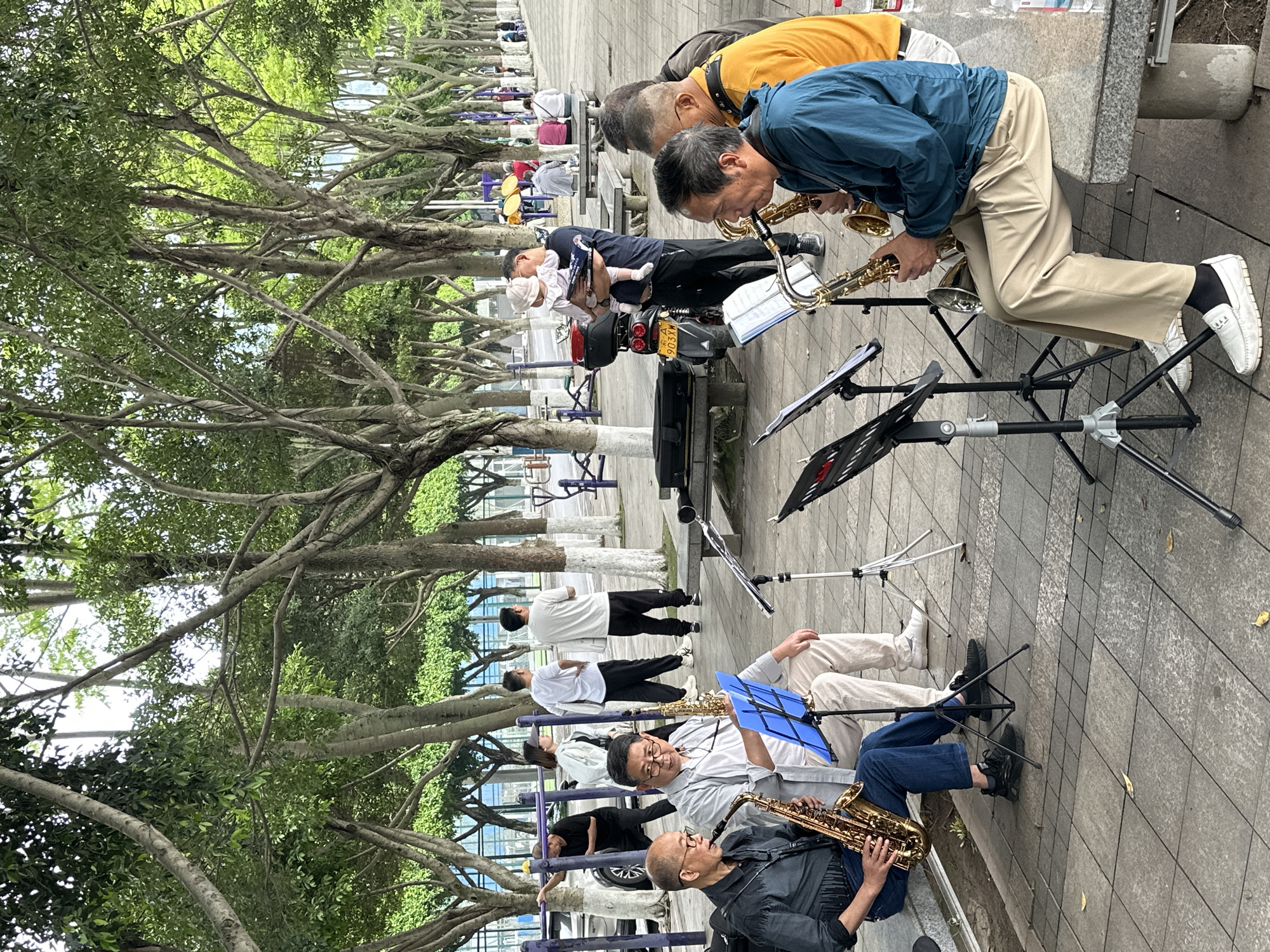In Jiangjin, China, a group of primarily elderly men are playing saxophones in a public square. Some of the men are sitting and others are standing. Each has a music stand holding sheet music. Several other people are in the background. The area is shaded by trees. There is a moped and a baby in the middle of the group.