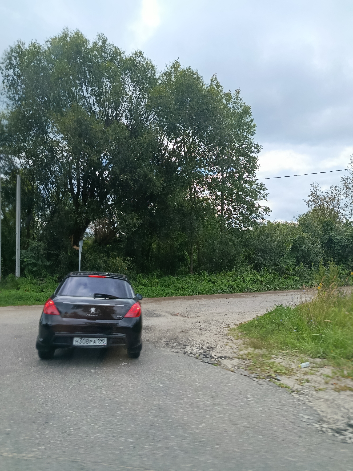 A black Peugeot hatchback is parked on a dirt road in a rural area. The car is facing away from the viewer, with its rear end visible. The license plate is partially obscured, but the visible portion reads "H308PA 190". The car is surrounded by lush green vegetation, with tall trees in the background. The sky is overcast, with a few clouds visible. The scene appears to be a quiet, secluded location. The location is identified as Снопок Старый, Russia.