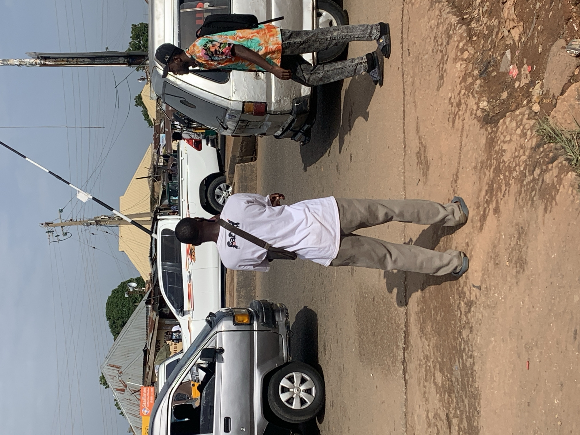 In Gura Top, Nigeria, a street scene unfolds under a bright sky. Two men stand on a dirt road next to a silver minivan. One man, wearing a white t-shirt and grey pants, faces away from the camera. The other man, in a colorful patterned shirt and distressed jeans, looks toward the minivan. Behind them, a white pickup truck is visible, with buildings and power lines in the background. A railway crossing barrier is angled across the frame. The ground is dusty, with some debris scattered around.
