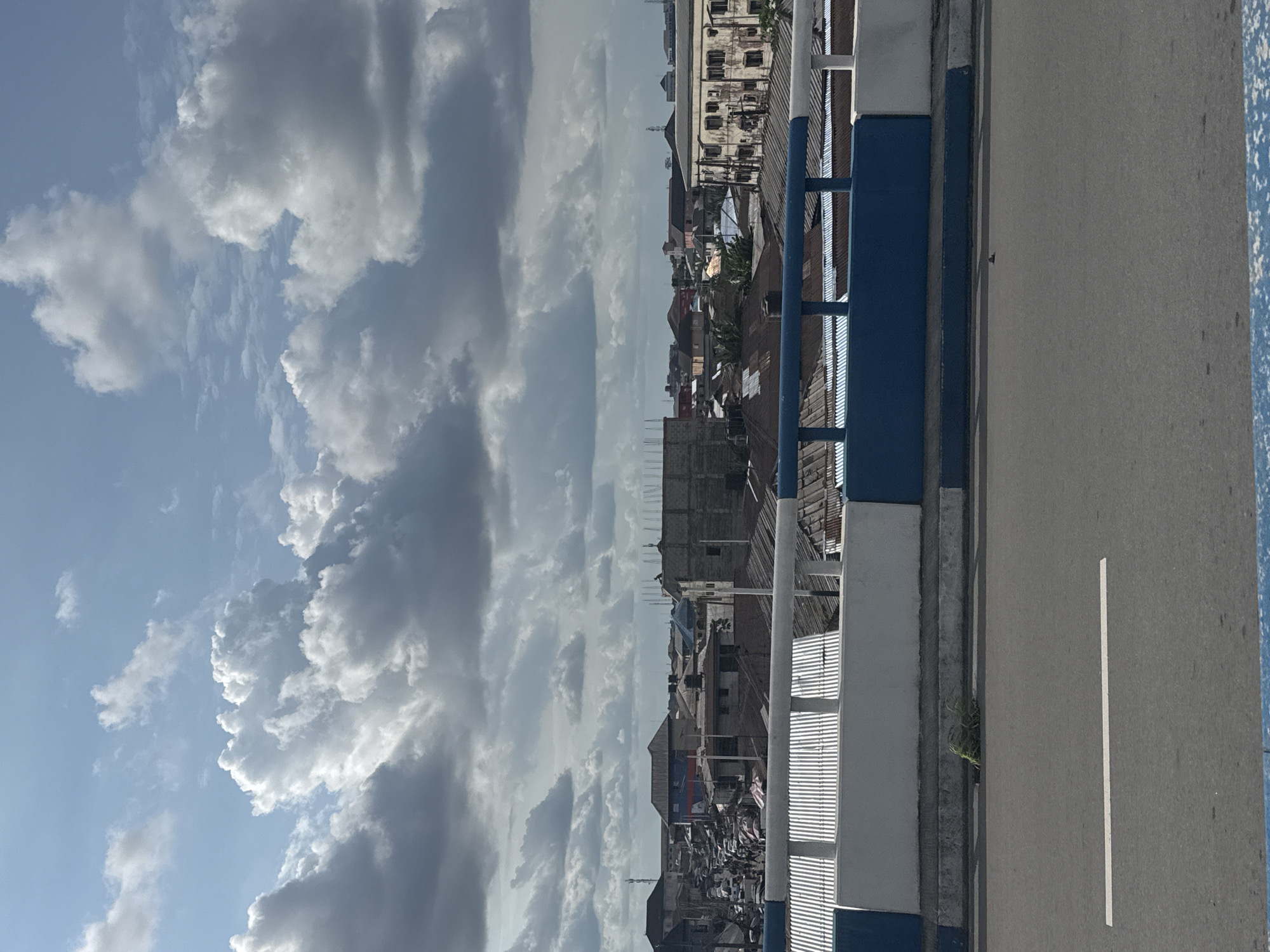 The image depicts a view from a bridge in what appears to be a densely populated area, likely within the city of Port Harcourt, Nigeria. The shot includes a section of the bridge's road surface with a single painted white lane marker. A barrier with a blue and white painted metal railing is visible on one side of the road. Beyond the railing, a crowded urban landscape can be seen, with buildings of varying heights and construction materials. The buildings are situated relatively close together. The sky is prominently featured, showing a mix of blue sky and heavy, cumulonimbus clouds. No people are directly visible in the frame, although the density of the settlement suggests a significant population presence.