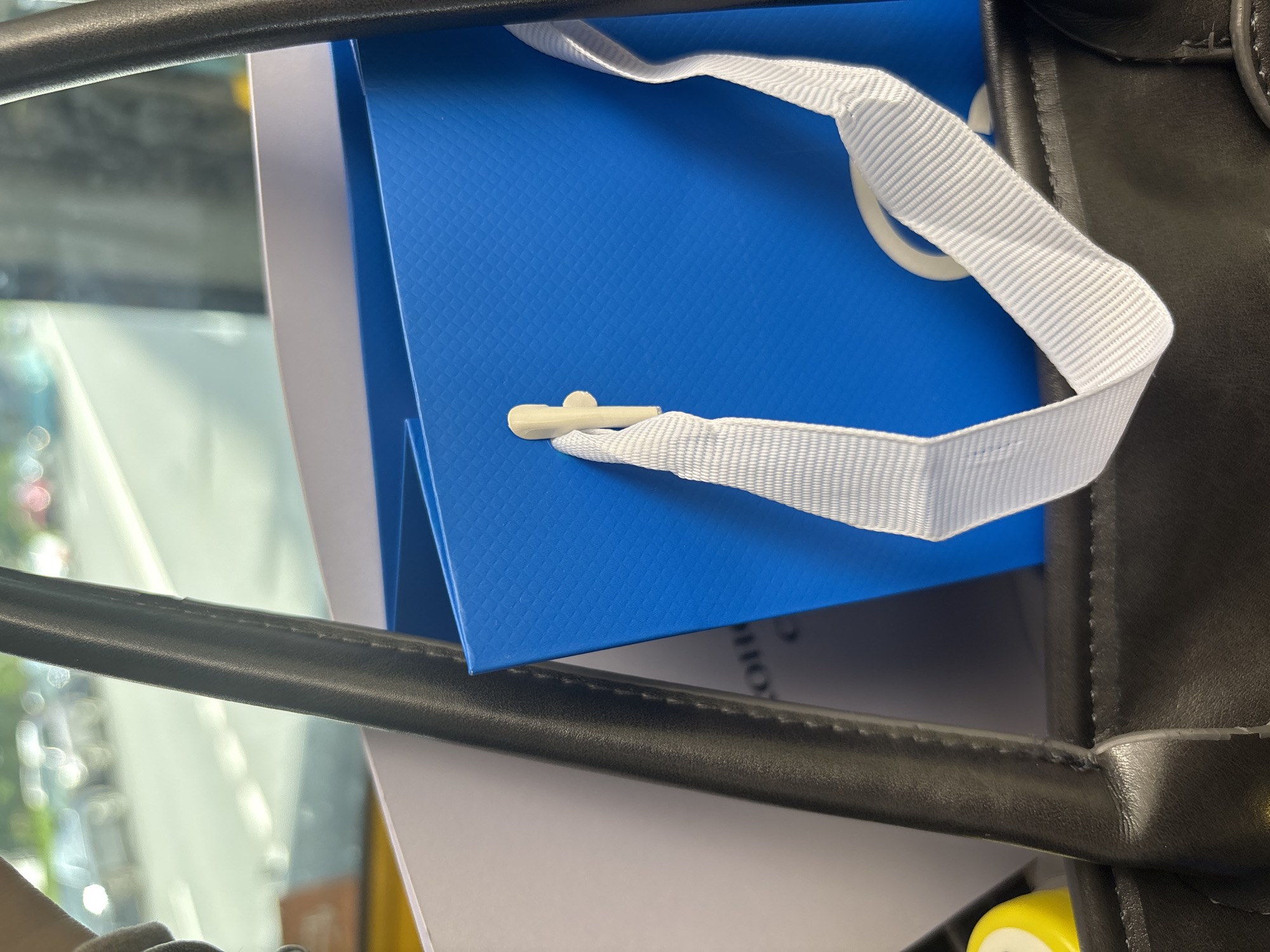 The image shows a close-up view of the interior of a black tote bag. A bright blue paper gift bag with a white ribbon handle is prominently featured inside the tote bag. The blue bag has a textured surface and a small, light-colored plastic closure. To the left of the blue bag, part of a white document or folder is visible. The background is blurred, showing a street scene with vehicles and greenery, suggesting the tote bag is being carried outdoors in an urban environment. The image does not provide enough information to determine the specific location in Saint Petersburg, Russia, or to identify any people.