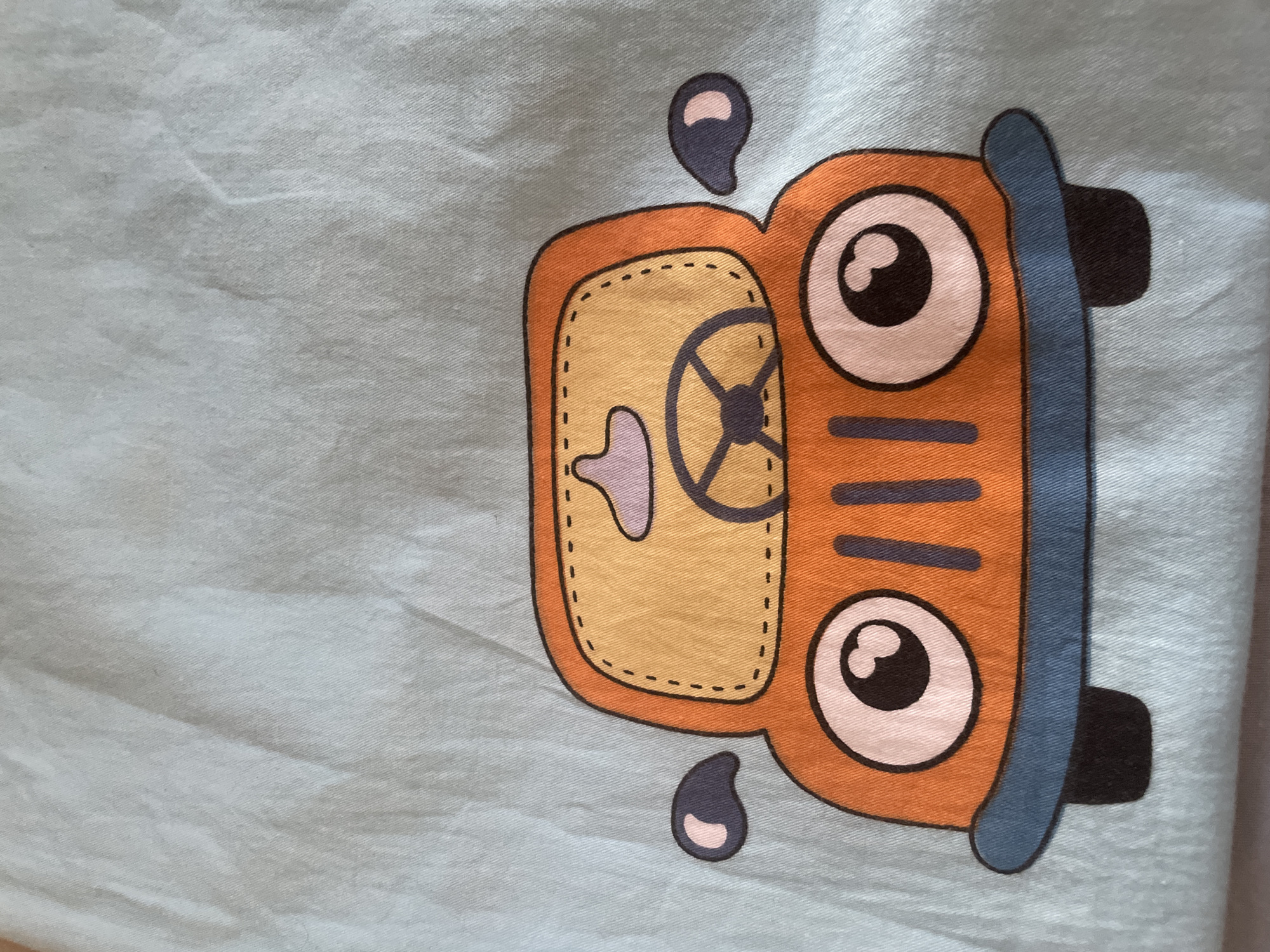 The image is a close-up photograph of a printed graphic of a cartoon orange car on a light blue fabric. The car has large, round eyes with black pupils, a black steering wheel, and a blue bumper. The fabric appears to be slightly creased and textured. There are no people or any other identifiable objects or environmental features in the image. The location in Tashkent, Uzbekistan cannot be determined from the provided image.