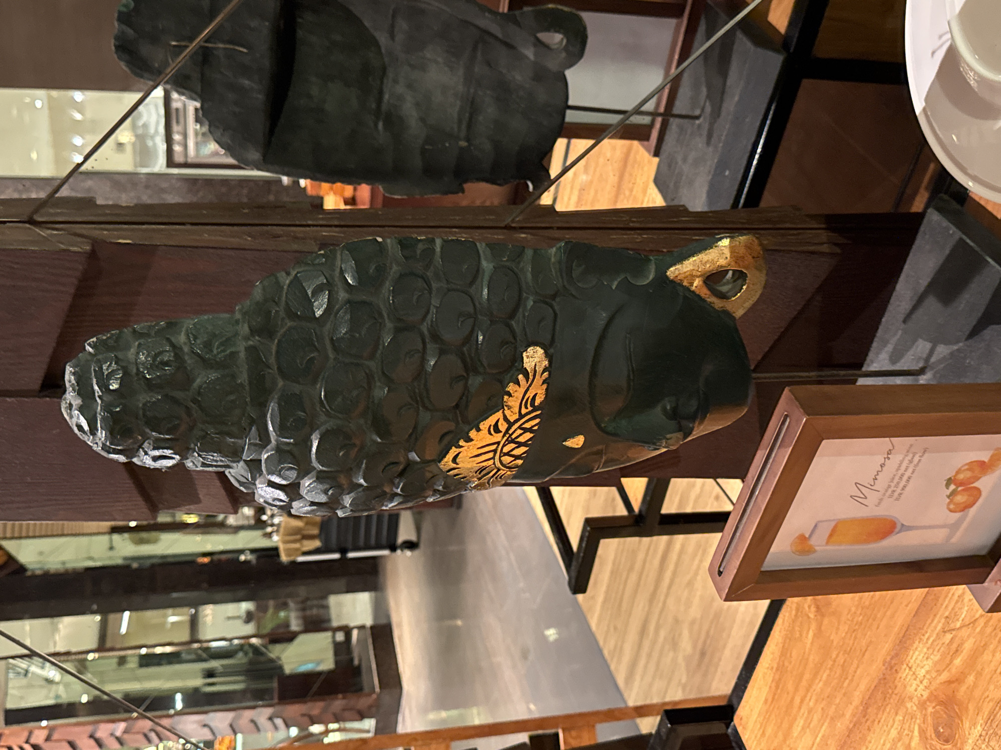 A decorative dark green carved wooden mask with gold accents is displayed indoors. The mask is carved to resemble a face with an ornate headdress, and a golden butterfly-like design is painted on the cheek. The mask is mounted on a dark metal stand and is positioned in front of a large dark wooden pillar. To the left of the mask, a small framed menu card displays an illustration of a mimosa cocktail and text describing it. The background shows a blurred interior with polished floors, reflective surfaces, and ambient lighting, suggesting a hotel lobby or restaurant setting. The overall scene is well-lit and appears to be a display of cultural or artistic elements. The presence of a menu card and display elements suggests the location is likely a food and beverage establishment in Kuta Selatan, Indonesia.