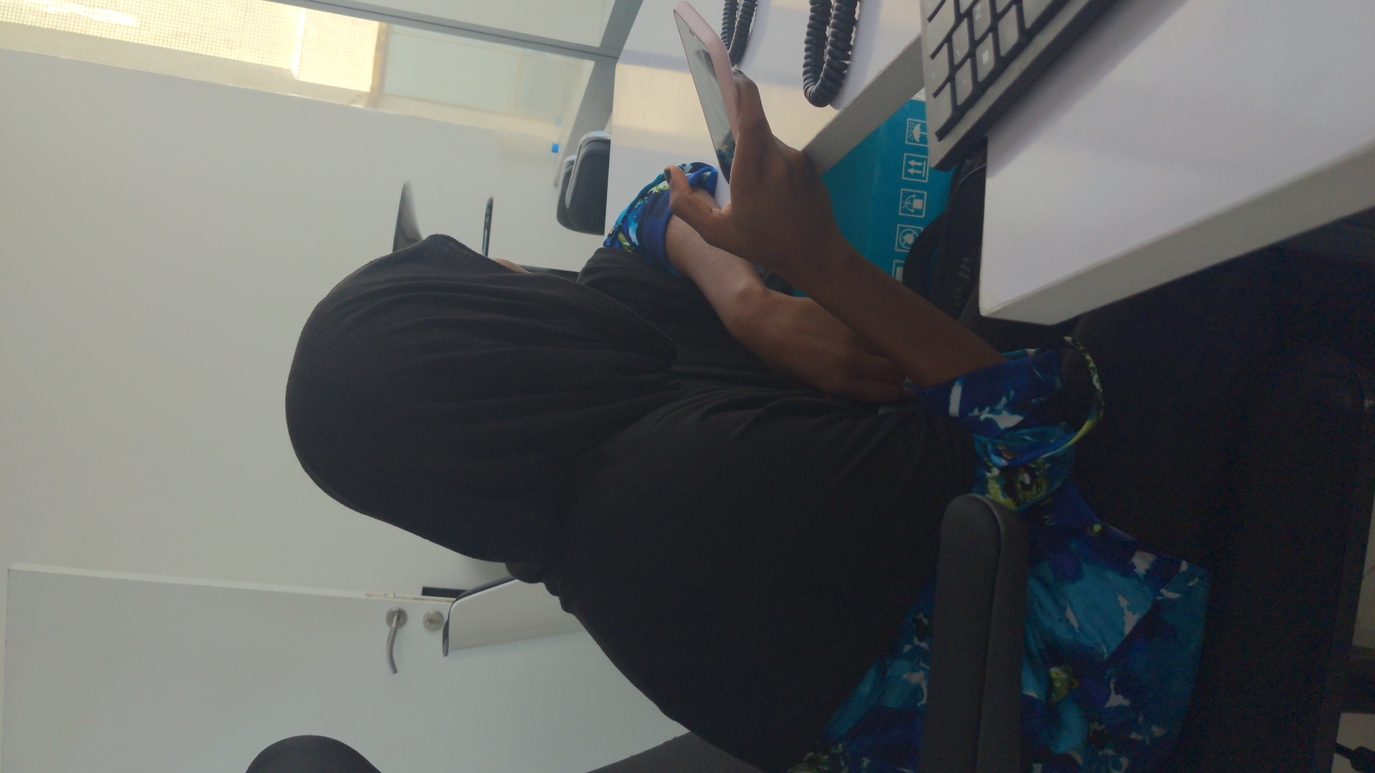 The image depicts an individual seated at an office desk in what appears to be an indoor office or workstation in Abuja, Nigeria. The person, who has a dark skin tone, is dressed in a black, long-sleeved garment, possibly an abaya or a similar modest dress, complemented by a black head covering. A vibrant blue, green, and white patterned sleeve is visible on their right arm.
The individual is actively engaged with a light-colored smartphone, holding it in both hands and looking down at its screen. Their posture suggests focused attention or relaxation while using the device.
The immediate surroundings include a white desk or table in the foreground. Partially visible on the desk are a black computer keyboard and a coiled black telephone cord. Beneath the desk, a light blue box adorned with international handling symbols, such as "this way up," "fragile," and "keep dry," can be seen. The person is seated in a dark office chair with a gray armrest. The background features plain white walls, and a bright light panel or window is visible at the top, suggesting well-lit conditions, likely daytime. Another white surface with small silver handles or hooks is present to the left of the frame. The image is captured from a slightly low angle, looking upwards towards the person and the desk.