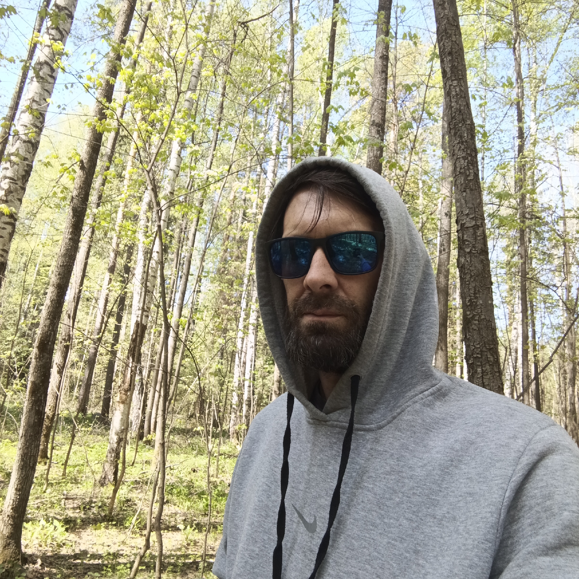 A man with a beard and sunglasses, wearing a gray hooded sweatshirt with a Nike logo, stands in a forest. The trees are mostly thin, with some showing white bark characteristic of birch trees. There is green vegetation on the forest floor. The sky is visible through the branches. The location is Markovo, Russia.