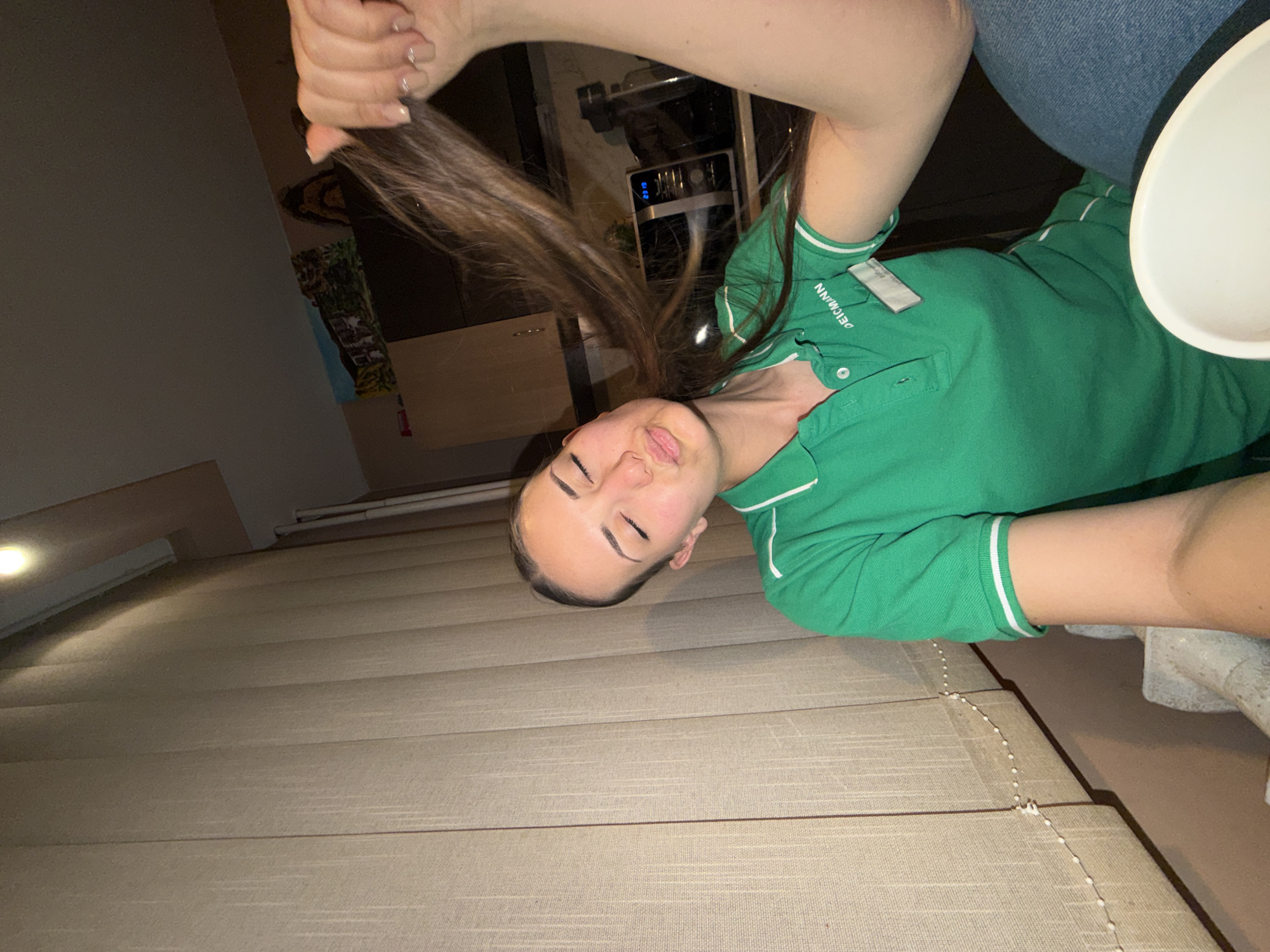A young woman in a green polo shirt is angled with her head towards the lower portion of the image. She's holding a strand of her brown hair. The background appears to be a kitchen environment. The location is unknown; there is no visual evidence of Buhovo, Bulgaria in the image. The subject is posing indoors. The ground beneath her head has corduroy lines.
