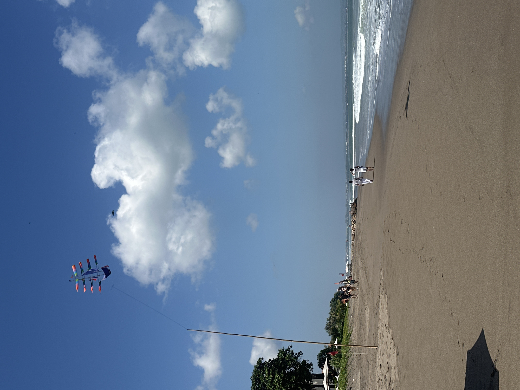 The image shows a beach in North Kuta, Indonesia, on a clear, sunny day. A large, colorful kite resembling a ship is flying high in the bright blue sky, tethered to a tall wooden pole on the sandy beach. The beach itself is wide and dark, with gentle waves breaking on the shore. In the distance, a few people are walking along the beach, and there are some palm trees and buildings visible beyond the sand. The overall atmosphere is peaceful and serene, with the kite serving as a focal point against the vast expanse of the sky and sea.