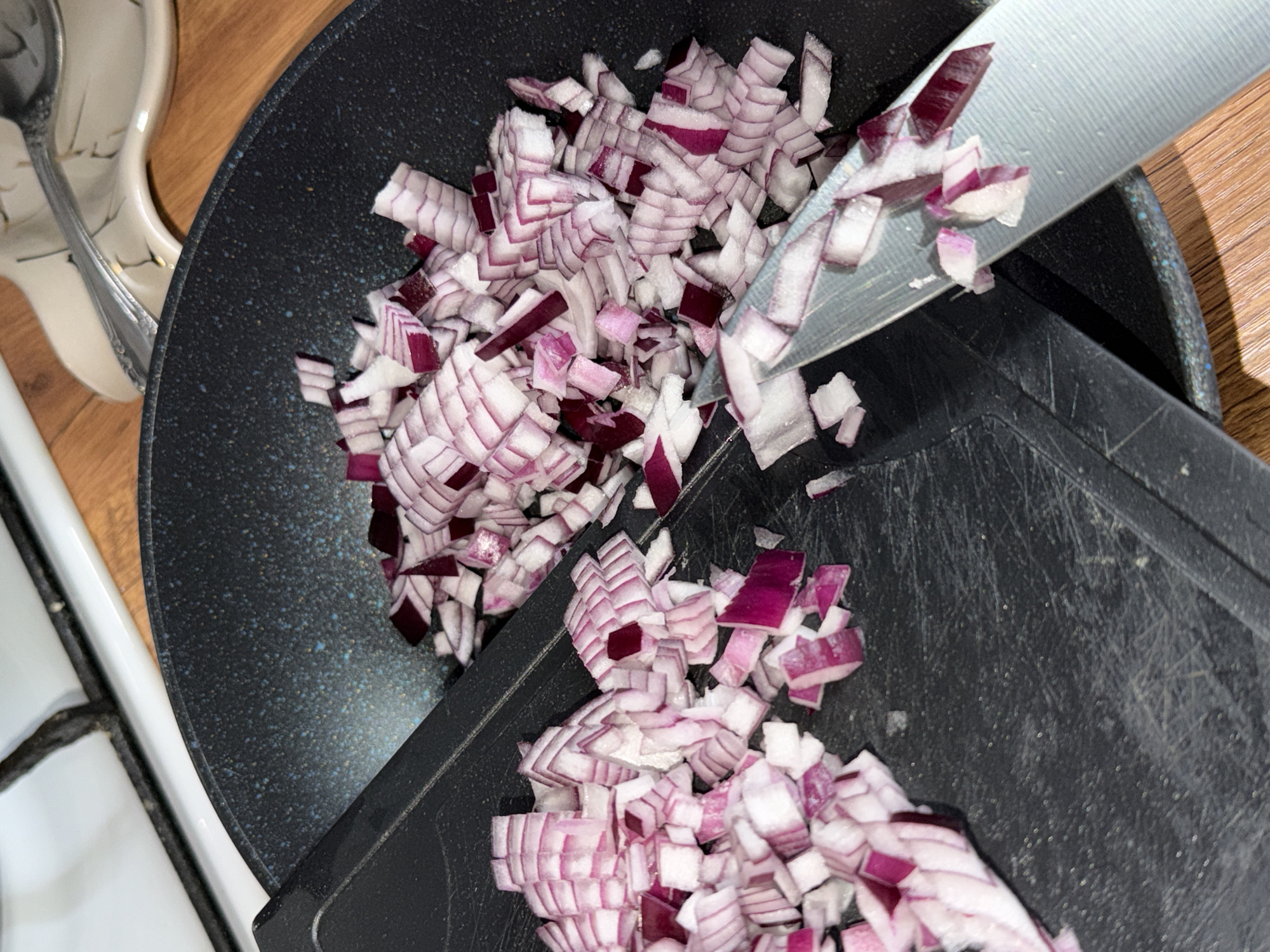 The scene is an indoor kitchen environment in Reshetnikovo, Russia. The image prominently features a quantity of diced red onions, characterized by their distinct white and purple layers. These chopped onions are distributed across a dark, worn cutting board and within a black, speckled non-stick frying pan. A silver-colored kitchen knife lies diagonally, partially on the cutting board and extending towards the pan, with several onion pieces resting on its blade. In the upper background, a section of a white stovetop with a dark burner grate is visible. To the right, a light-colored, patterned spoon rest holds a metal spoon. All elements are situated on a light brown wooden surface.