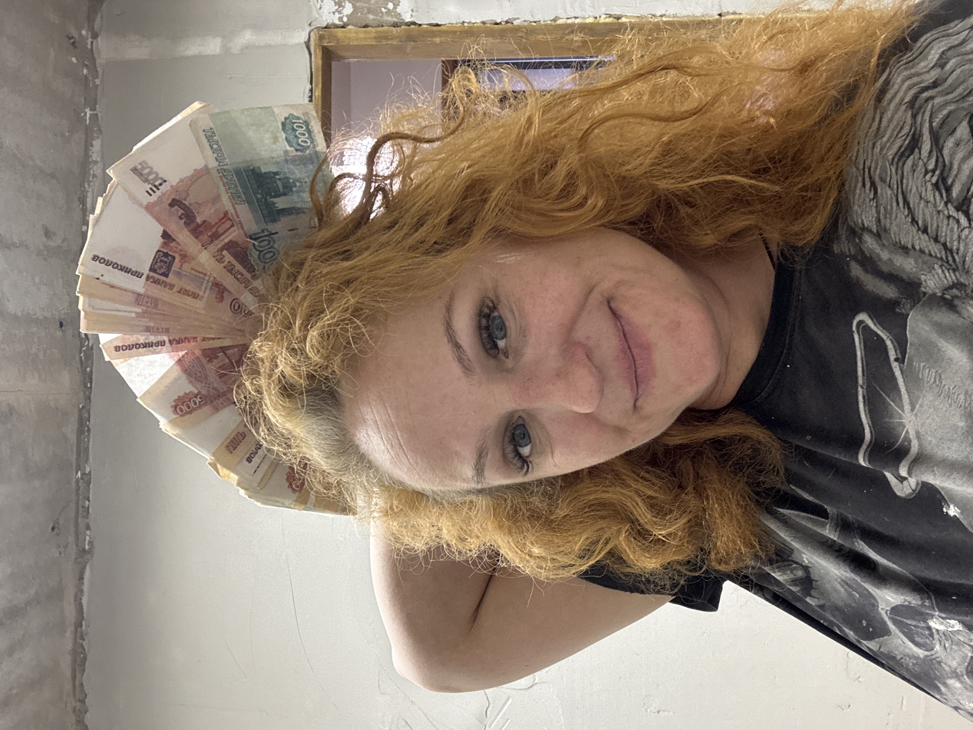 An image, captured at a tilted angle, shows a woman with long, curly red hair and blue eyes smiling subtly towards the camera. She is positioned indoors, likely in Chaikovsky, Russia. She wears a dark t-shirt adorned with a prominent grey and white graphic design, possibly depicting a wolf or a similar creature.
Around her head, she holds a fan of banknotes, playfully arranged like a crown. The visible currency includes Russian rubles of various denominations, such as 1000 (green), 5000 (reddish-pink), and 100. Key text on the banknotes includes "1000" and "ТЫСЯЧА РУБЛЕЙ" (thousand rubles), and "5000" and "ПЯТЬ ТЫСЯЧ" (five thousand rubles). Notably, several of the notes also clearly display "БИЛЕТ БАНКА ПРИКОЛОВ" (joke bank ticket), indicating they are novelty or fake currency.
The background features unfinished or renovating walls, with exposed concrete or plaster showing a rough texture. An unpainted, golden-hued wooden window frame is visible in the upper right portion of the image. The overall setting suggests a room undergoing construction or refurbishment. The woman's expression is one of slight amusement or confidence.