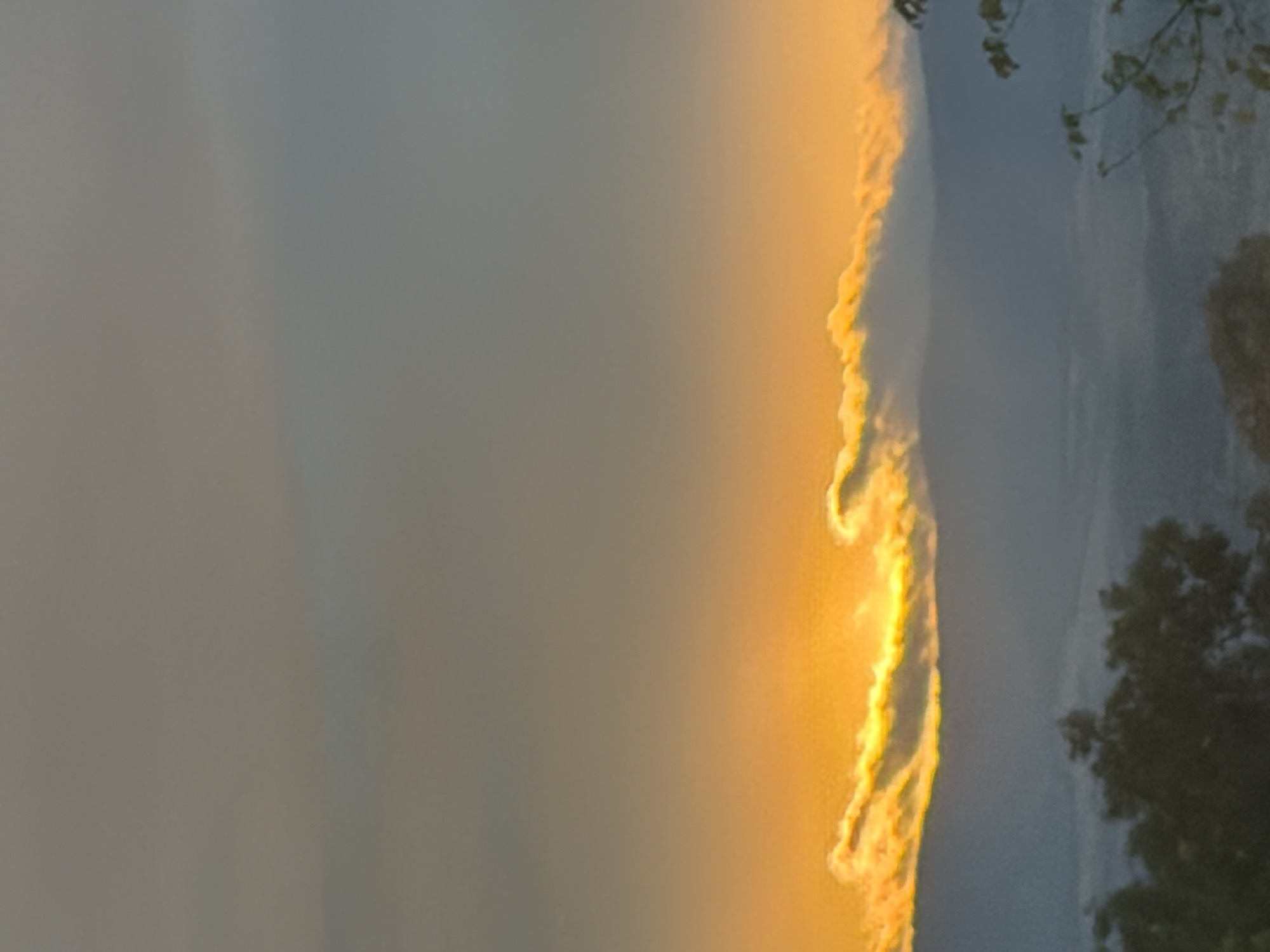 The image depicts a misty mountain landscape at sunrise. The sun is low on the horizon, casting a warm orange and yellow glow on the clouds that are scattered across the sky. The mountains in the background are shrouded in a soft haze, with layers of mist creating a sense of depth. In the foreground, the silhouette of trees is visible, adding a touch of darkness to the scene. The overall impression is one of tranquility and natural beauty.
This image was taken in Blagoevgrad, Bulgaria, a city known for its mountainous surroundings. The specific location is likely a viewpoint overlooking the city and the Rila Mountains.