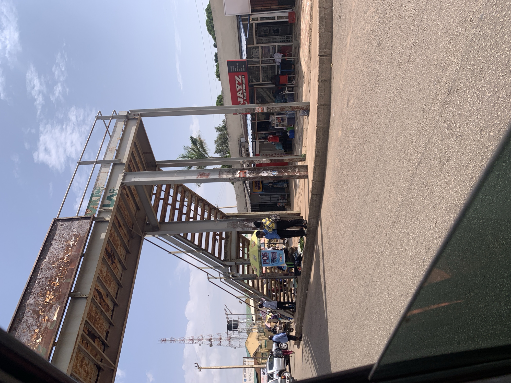 The media file captures a street scene in Jos, Nigeria, under a bright, sunny sky with scattered clouds. The orientation of the image is vertical. In the foreground, a grey, textured asphalt road with a concrete curb runs along the right side. To the left of the road, a large, somewhat dilapidated pedestrian overpass structure dominates the scene. The overpass is made of weathered metal beams and planks, showing signs of rust and peeling paint, particularly on the signage attached to its side.
Beneath the overpass, several people are visible, some appearing to be vendors with their goods displayed under a yellow umbrella. There are also shoppers and passersby. The activity seems to be focused around the stalls and the entrance to the overpass.
Behind the overpass and the people, a row of shops lines the street. One shop clearly displays the sign "JAYZ CONFECTIONERIES" in large red letters. Other shops have awnings and various goods visible through their windows or displays. A palm tree is visible to the left of the shops. Further in the background, a telecommunications tower stands tall, and the roofs of buildings suggest a populated urban environment.
A white pickup truck is parked on the road, and another vehicle is partially visible. The overall impression is that of a busy street scene, likely during daytime, with commerce and pedestrian traffic taking place around a functional but aged piece of infrastructure. The lighting suggests it is midday, with clear, bright sunlight casting shadows.