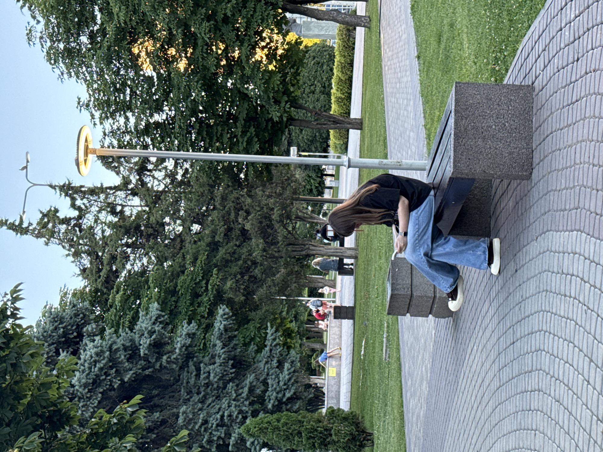 A young woman with long, brown hair, wearing a black t-shirt and blue jeans, sits on a granite bench in a park. She is looking down at her mobile phone. Her legs are crossed, and she is wearing white sneakers with black accents. The bench is located on a paved area with a circular pattern. In the background, several trees, including a tall, slender evergreen and mature deciduous trees, create a lush green canopy. A modern lamppost with a circular light fixture stands to the left of the woman. Further in the background, other people are visible, some walking and others sitting on a low stone wall. The setting appears to be a public park in Rostov-on-Don, Russia.