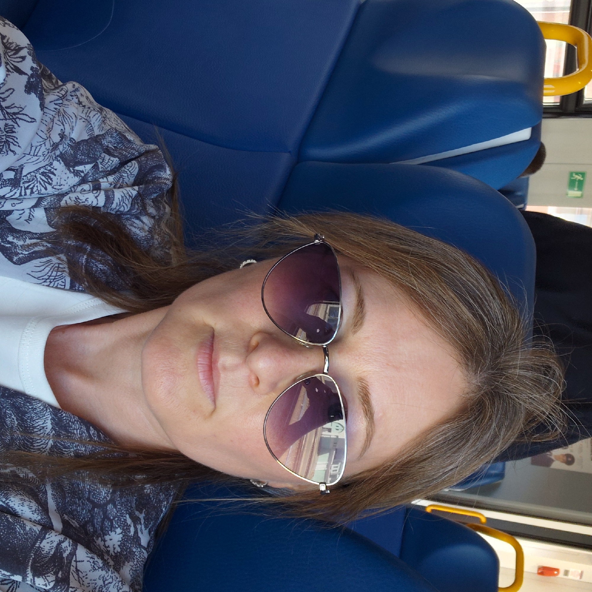 This is a close-up, rotated image of a woman wearing sunglasses, lying on a blue seat, possibly on public transport. She has medium-length, brown hair with some gray strands. Her skin appears fair. She is wearing a white top and a patterned garment with a black and white design, possibly a blouse or shirt. The sunglasses have dark, reflective lenses. The background consists of blue seats and a window with a yellow handle. A green sticker with a white symbol is visible on the window frame. The scene appears to be indoors, with natural light coming from the window, suggesting it could be daytime. There is no clear indication of the weather. The woman's eyes are not visible due to the sunglasses, but her mouth is slightly open, giving a relaxed or perhaps sleepy expression. The overall impression is of someone resting during a journey.
