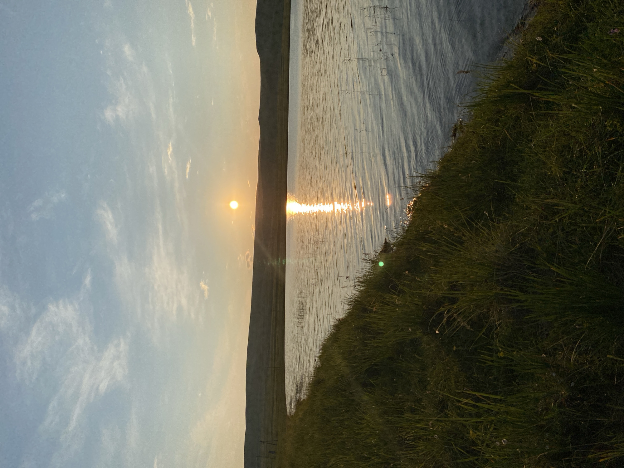 A serene landscape features a body of water reflecting the setting sun. The sun, a bright orb, is positioned above a distant, rolling hill. Its golden rays create a shimmering path across the rippling surface of the water. The foreground is dominated by a grassy bank, with tall green blades of grass swaying gently. The sky above is a soft blend of blue and pale orange, with wispy white clouds scattered across it. There are no people or animals visible in the frame. The scene conveys a sense of tranquility and natural beauty.