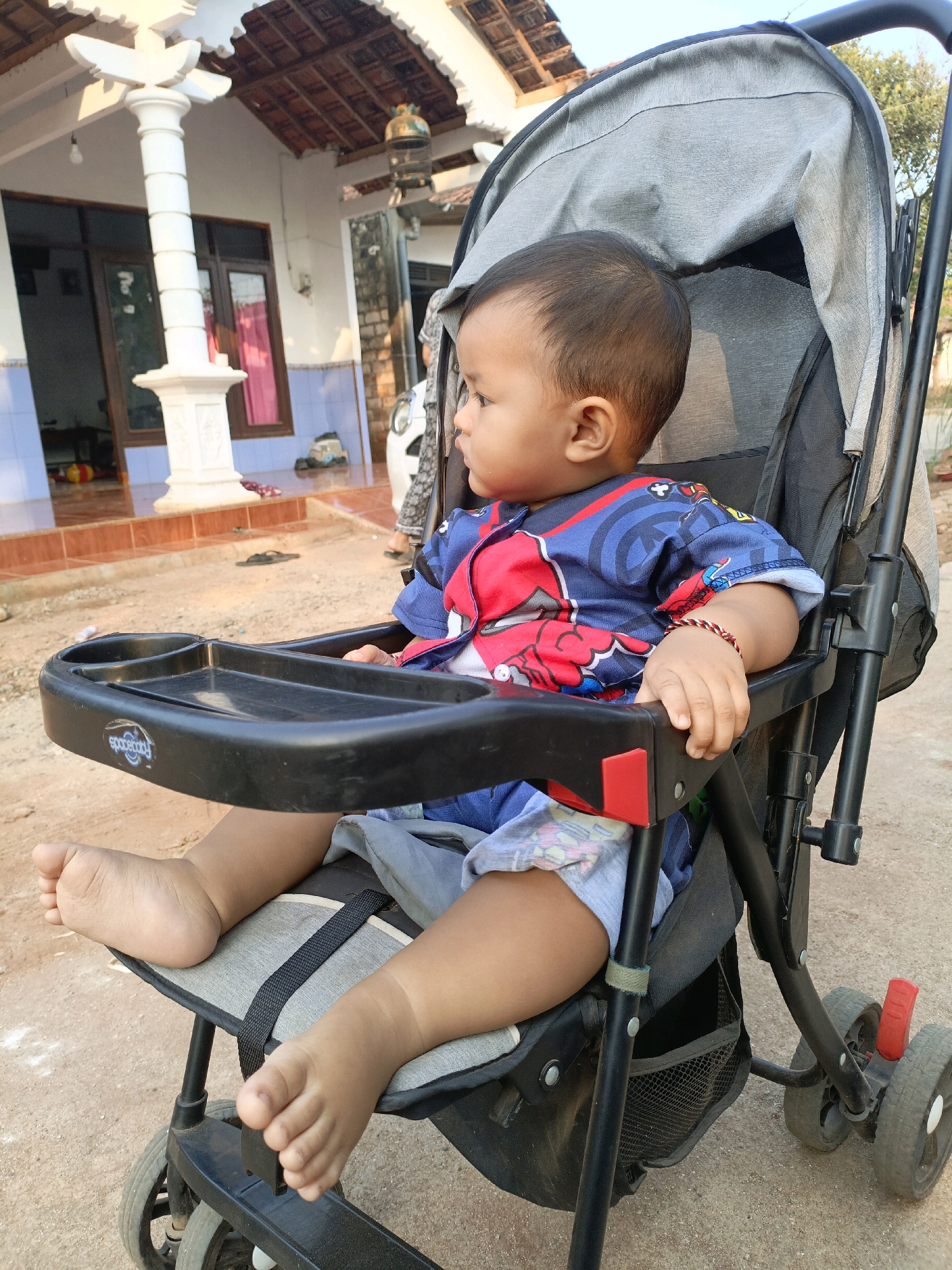 A young child sits in a stroller outdoors in Jenu, Indonesia. The child, facing to the left, wears a blue and red shirt with a graphic print and light blue shorts. A red and white braided bracelet is on the child's left wrist. The stroller is dark gray with a black tray in front of the child. The background shows a house with a tiled roof, white pillars, and a porch. The ground is dirt or gravel. The lighting suggests it is daytime.