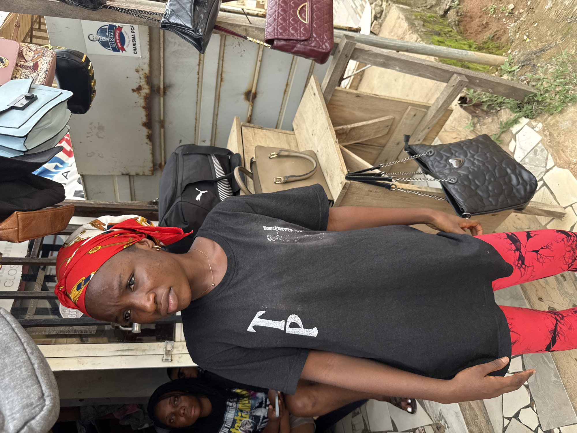 The image features a young woman wearing a black t-shirt and red patterned leggings, with a red and white headscarf tied around her head. She is looking at the camera with a neutral expression. The background suggests an outdoor market stall with various bags displayed for sale. Visible bags include a light blue handbag, a black backpack, a tan handbag, and a black patterned handbag with a chain strap. There are also some wooden crates and a metal structure in the background. To the left, there's a partial view of a gray bag and another person in black clothing. Further back, another person is visible wearing a black t-shirt with text on it.
A poster with text is visible in the background. The text reads "GLOBAL" and "CHARISMA PGF".
The scene appears to be set during daylight hours. The overall impression is of a vibrant market environment, likely in Abuja, Nigeria, given the context.