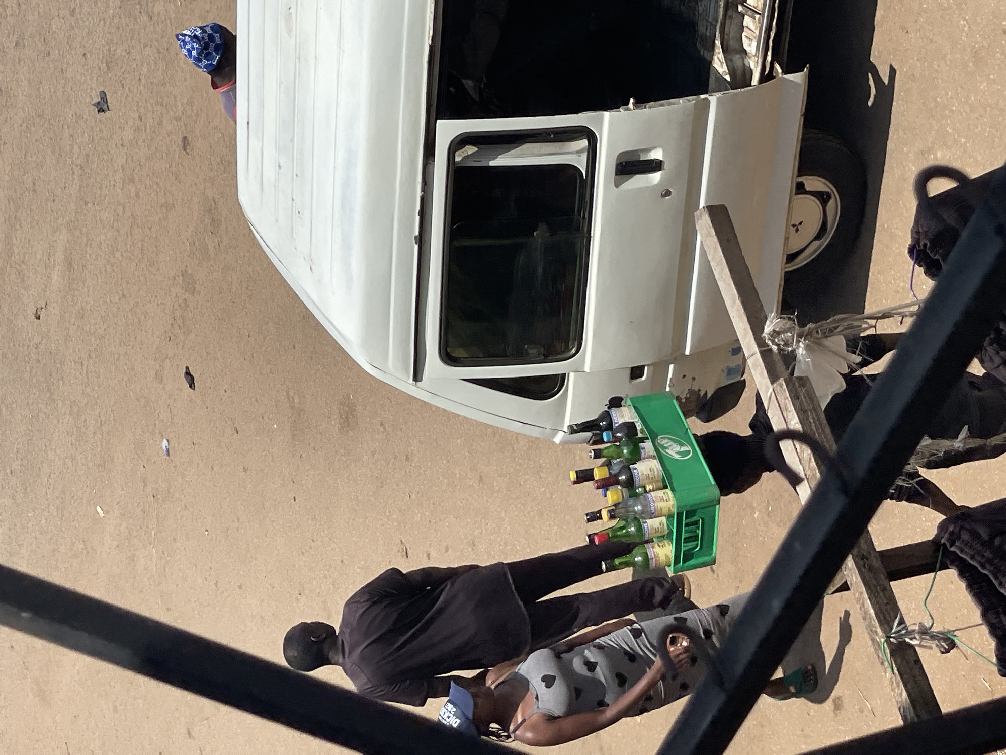 In an outdoor urban environment, likely on a dirt road, a white van with its side door open is parked. A woman wearing a gray dress with a heart pattern and a blue baseball cap walks towards the van. She is carrying a cell phone in her right hand. Next to her, a man in a black t-shirt and dark pants stands facing away from the camera. Another person, partially obscured, is carrying a green crate filled with bottles on their head, walking towards the van. The crate contains an assortment of bottles, some appearing to be alcoholic beverages and others labeled with text that suggests they might be medicinal. In the background, a person wearing a blue and white patterned hat is visible near the van. The ground is a light brown, dusty surface with some scattered debris. The foreground is dominated by dark, angled metal bars, suggesting the photo was taken from an elevated position.