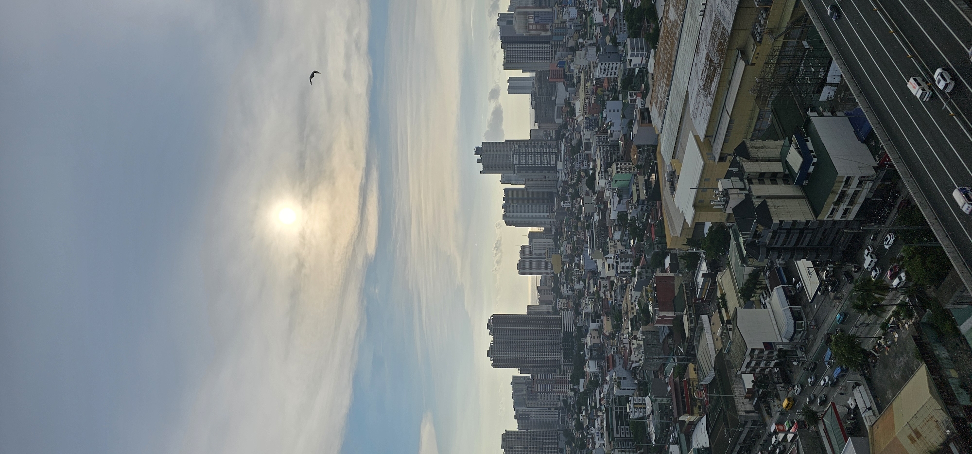This is a vertical, high-angle, full shot of a cityscape in Makati, Philippines, at what appears to be dawn or dusk. The sun is visible in the upper center of the frame, casting a soft glow through hazy clouds. A solitary bird is in flight in the upper right quadrant. The mid-ground is dominated by densely packed, multi-story buildings of varying architectural styles and ages, with some modern high-rises interspersed with older, more traditional structures.
The lower portion of the image shows a section of a multi-lane highway with several vehicles traveling on it. The highway is elevated above the surrounding urban landscape, which includes residential and commercial buildings, trees, and a gas station. The overall impression is of a busy, sprawling metropolis with a clear contrast between older and newer developments. The lighting suggests a time of day with diffused sunlight, possibly indicating overcast conditions or early morning/late evening light.
