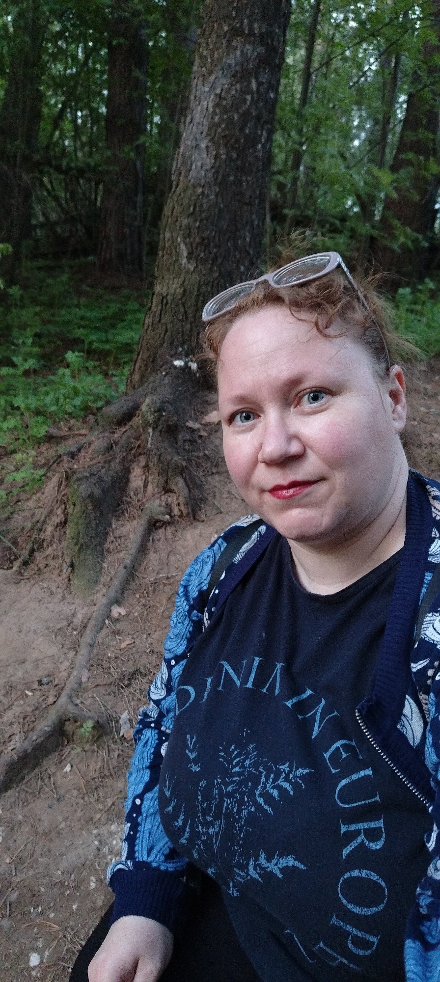 A fair-skinned woman with red hair and blue eyes is posing for a selfie outdoors. She wears a black T-shirt with the words "Journey in Europe" and a blue floral cardigan. Her glasses are perched on her head. Behind her is a large tree with exposed roots, and a forest is visible in the background. The location is Chaikovsky, Russia.
