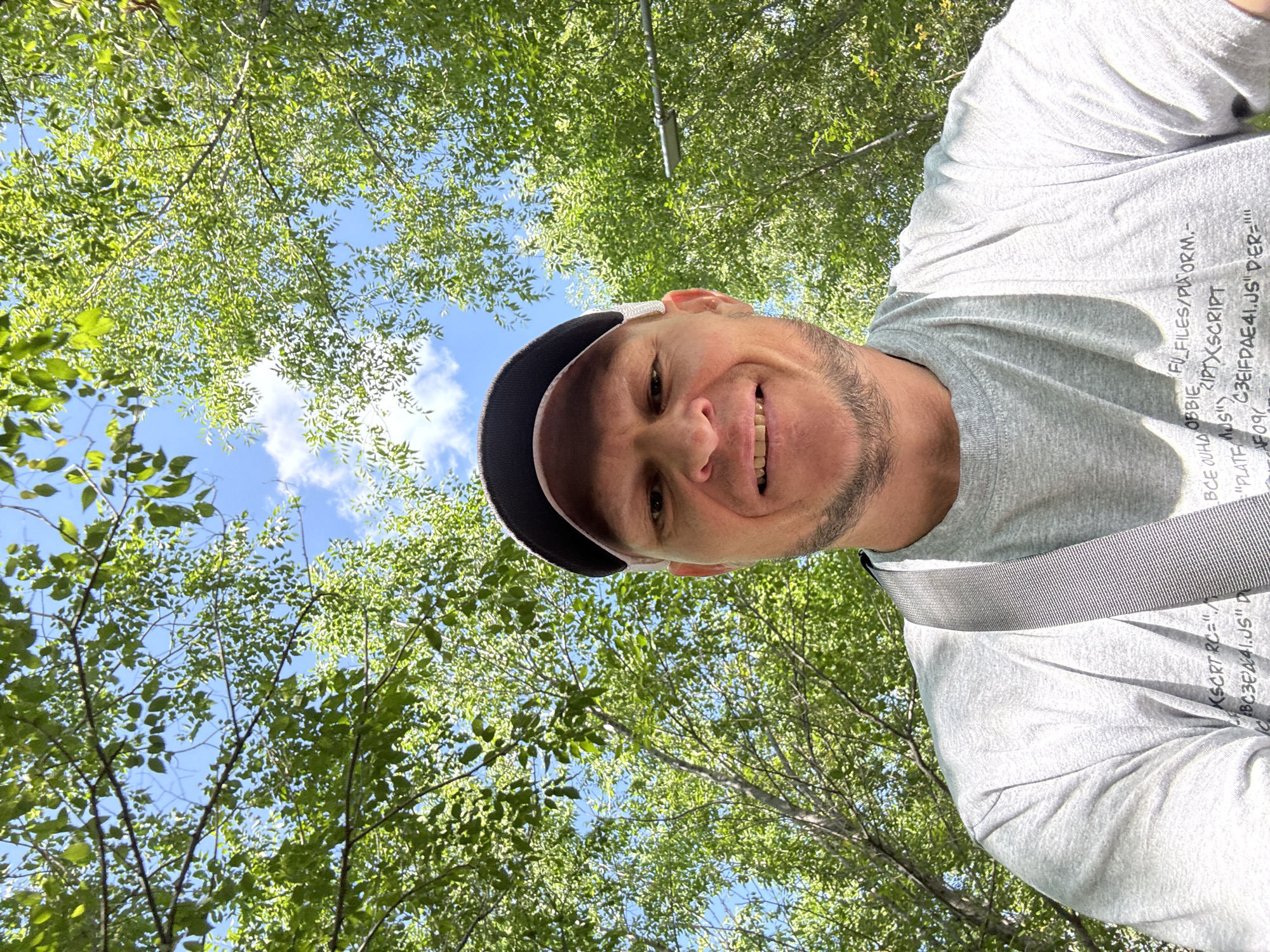 A man with a light complexion, a shaved head, and a goatee is taking a selfie outdoors. He is wearing a grey t-shirt with text printed on it, a dark baseball cap with a white brim, and a grey strap across his chest. He is smiling. The background consists of green trees and a blue sky with some clouds. The image is taken from a low angle, looking up at the man and the trees. There is a street light visible in the background. The location is identified as Oskemen, Kazakhstan.