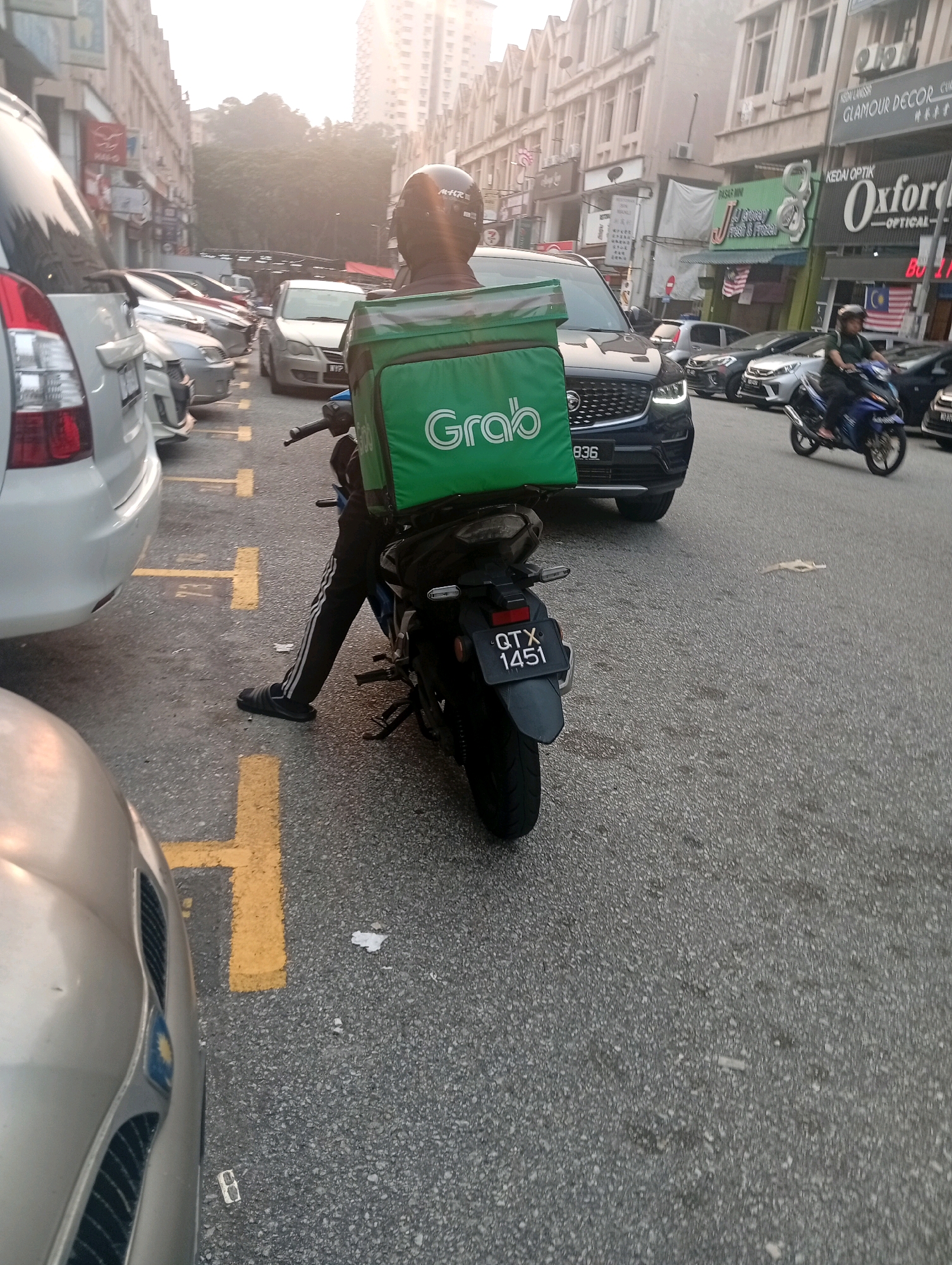 A medium-shot, eye-level, outdoor, daytime, street-level photograph captures a Grab delivery rider on a motorcycle parked on a street in what appears to be Selayang Municipal Council, Malaysia. The rider is wearing a black helmet and black pants with white stripes down the sides. They are seated on a black motorcycle with a large green Grab delivery box on the back. The motorcycle has a license plate reading "QTX 1451". The rider's left leg is extended forward, and they are wearing black slippers.
In the background, various cars are parked along the street, including a white SUV on the left and a dark blue SUV in the center. The street is paved with asphalt and marked with yellow parking lines. Buildings line the street, displaying shop signs, some of which are in Malaysian. The sky is hazy and bright, suggesting late afternoon or early morning. A few people are visible in the distance, including another motorcyclist on a blue bike further down the street. The overall scene depicts a typical urban street environment with commercial activity.