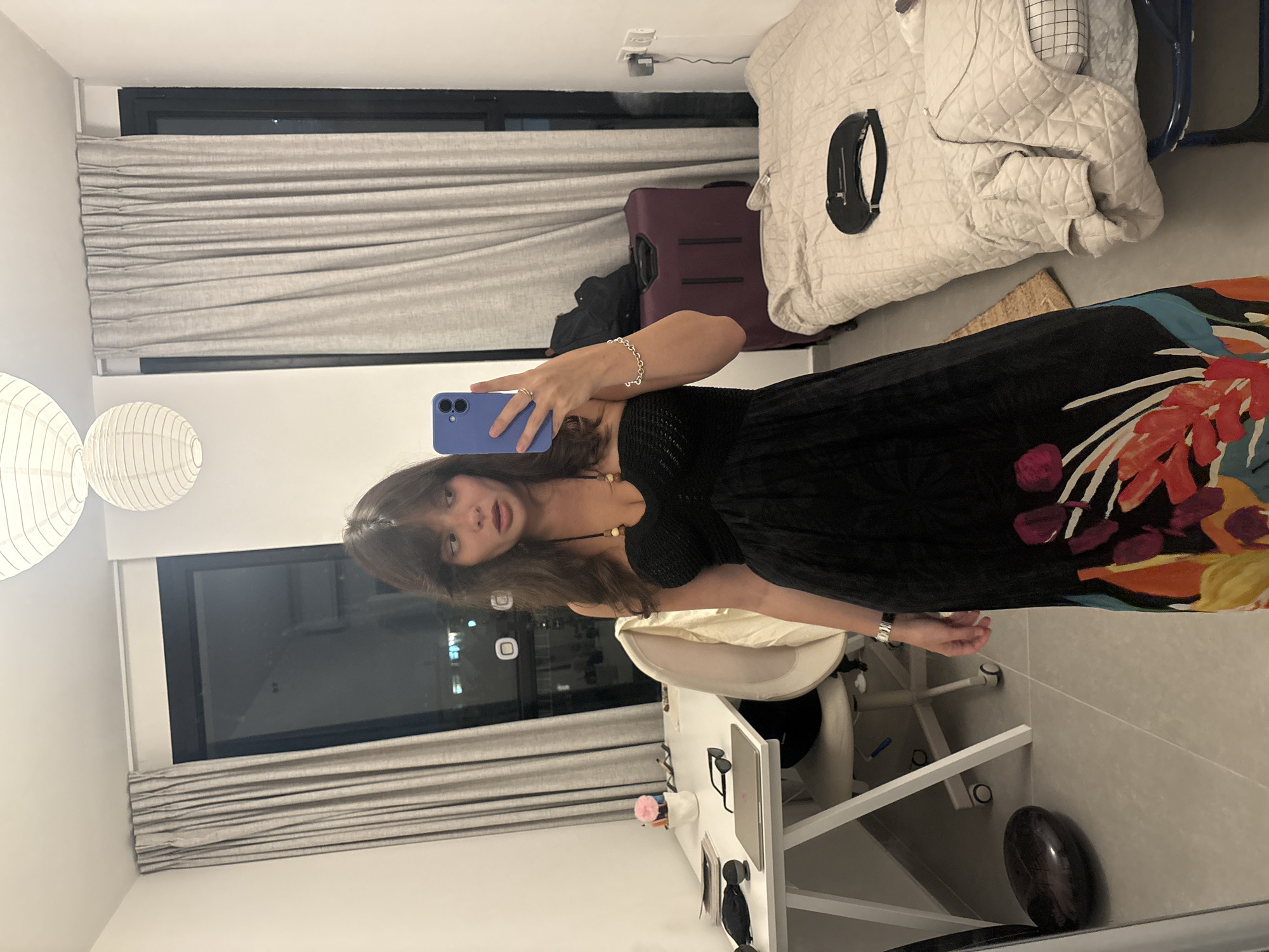A young woman stands in a bedroom, taking a mirror selfie with her phone. She is wearing a black strapless dress with a colorful floral pattern on the bottom. The room is minimalist, with a white desk and chair on the left side, and a bed with quilted bedding on the right. A large window with gray curtains dominates the background, offering a view of the city lights of Tel-Aviv at night. A dark purple suitcase and a black handbag are visible near the bed. The overall impression is of a contemporary and organized living space.