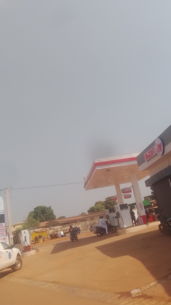 The scene depicts an outdoor view of an ORCA'S gas station located on reddish-brown dirt ground in Korhogo, Côte d'Ivoire, under a light gray sky. A prominent red and white canopy covers multiple white fuel pumps. A dark-colored building is attached to the right side of the gas station structure. On the far left, a white pickup truck, partially visible, displays a red cross-like emblem on its side and is positioned near a white fuel pump. At the pumps, an individual dressed in white is seated on a dark scooter, while another person stands near a different pump. A third individual on a dark scooter is observed moving across the mid-ground. In the background, various low-rise buildings and scattered trees are visible. Power lines extend horizontally across the scene.