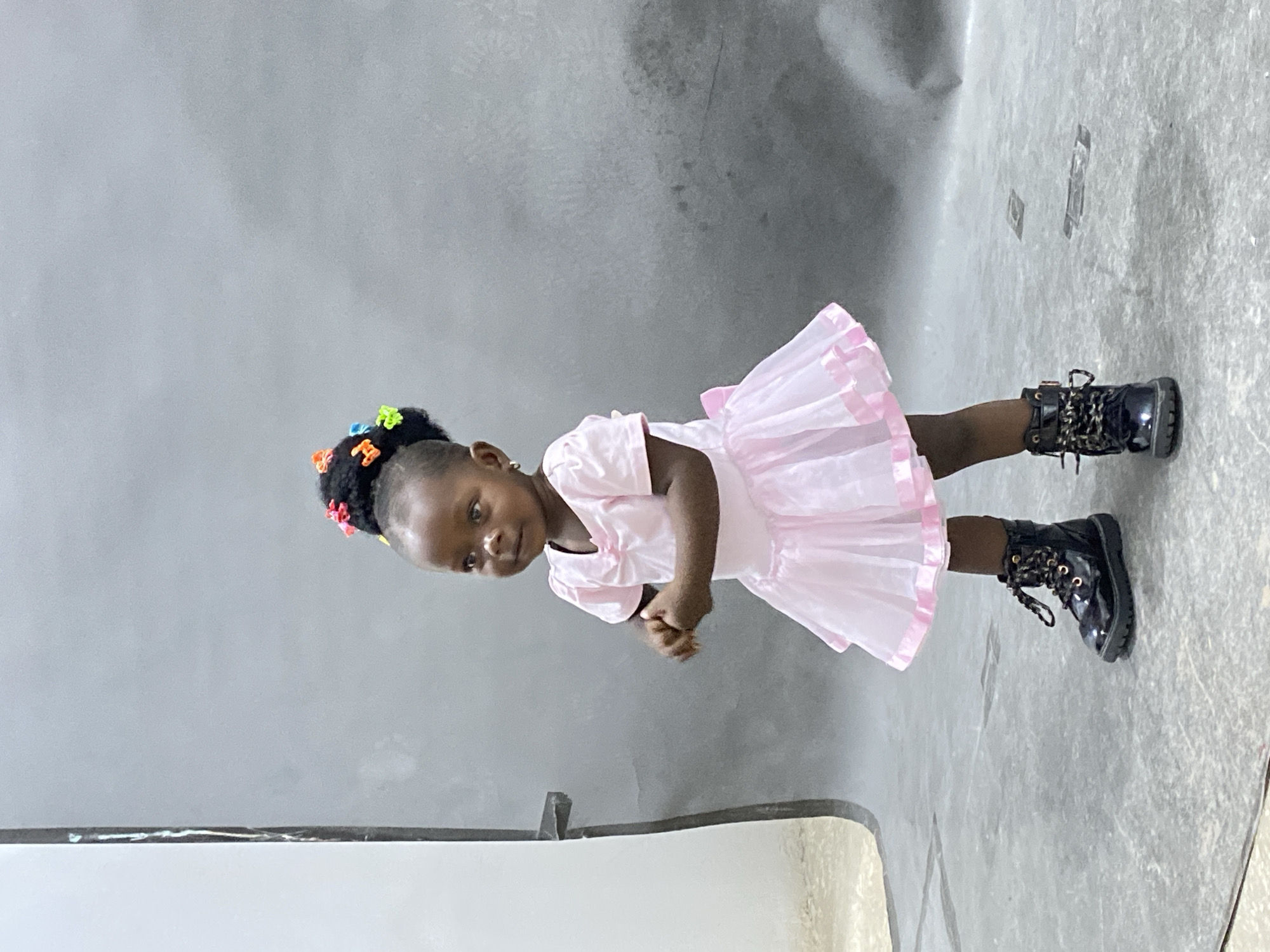 A young girl is standing in front of a light grey wall and a white, partially visible object. The girl has dark hair with colorful clips, and she wears a light pink dress and black, lace-up boots. She is looking at the camera. The location is Gura Top, Nigeria.