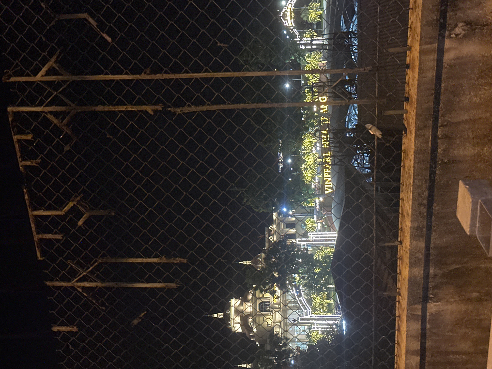 The image shows a night view through a wire fence of a building complex illuminated by numerous lights. The architecture of the buildings appears grand with domes and spires, reminiscent of a palace or resort. String lights are draped around trees and along pathways, creating a festive atmosphere. A prominent sign with the text "VINPEARL NHA TRANG" is visible in the foreground. The overall scene suggests a resort or entertainment complex, likely Vinpearl Land in Nha Trang, Vietnam, decorated for an event or evening illumination. The foreground shows a concrete barrier and more fencing, indicating the viewpoint is from outside the property.