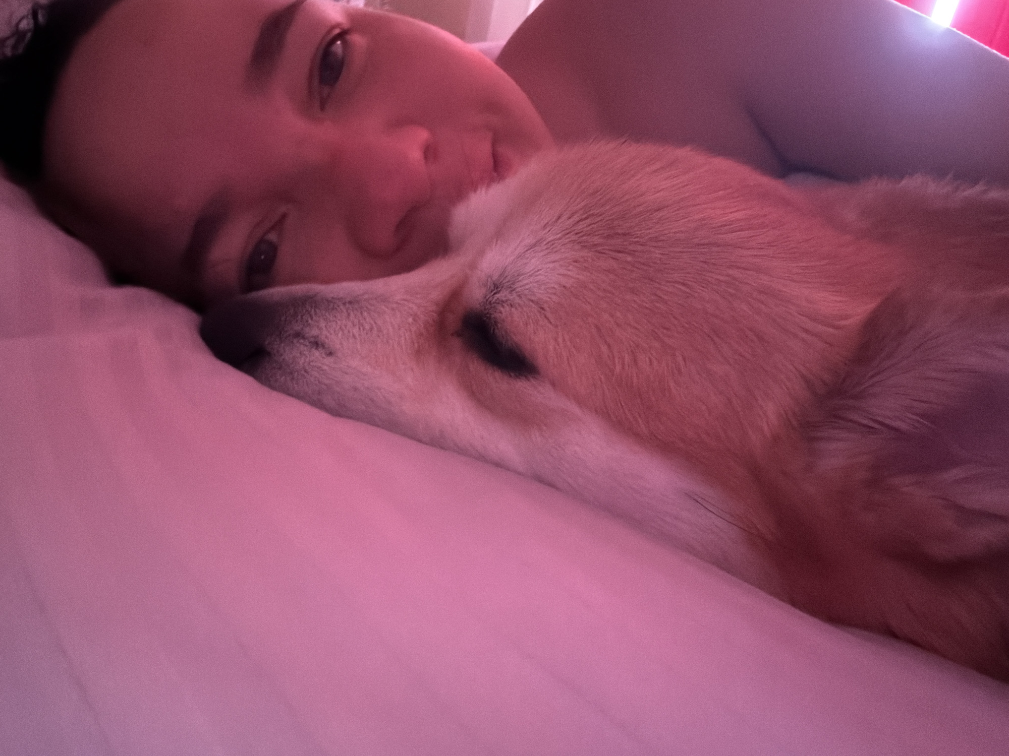 A person is lying down with a dog. The person's face is visible, and they appear to be looking at the camera. The dog is lying next to the person, and its head is resting on the pillow. The location is unknown. The city of Chaikovsky, Russia is not mentioned.