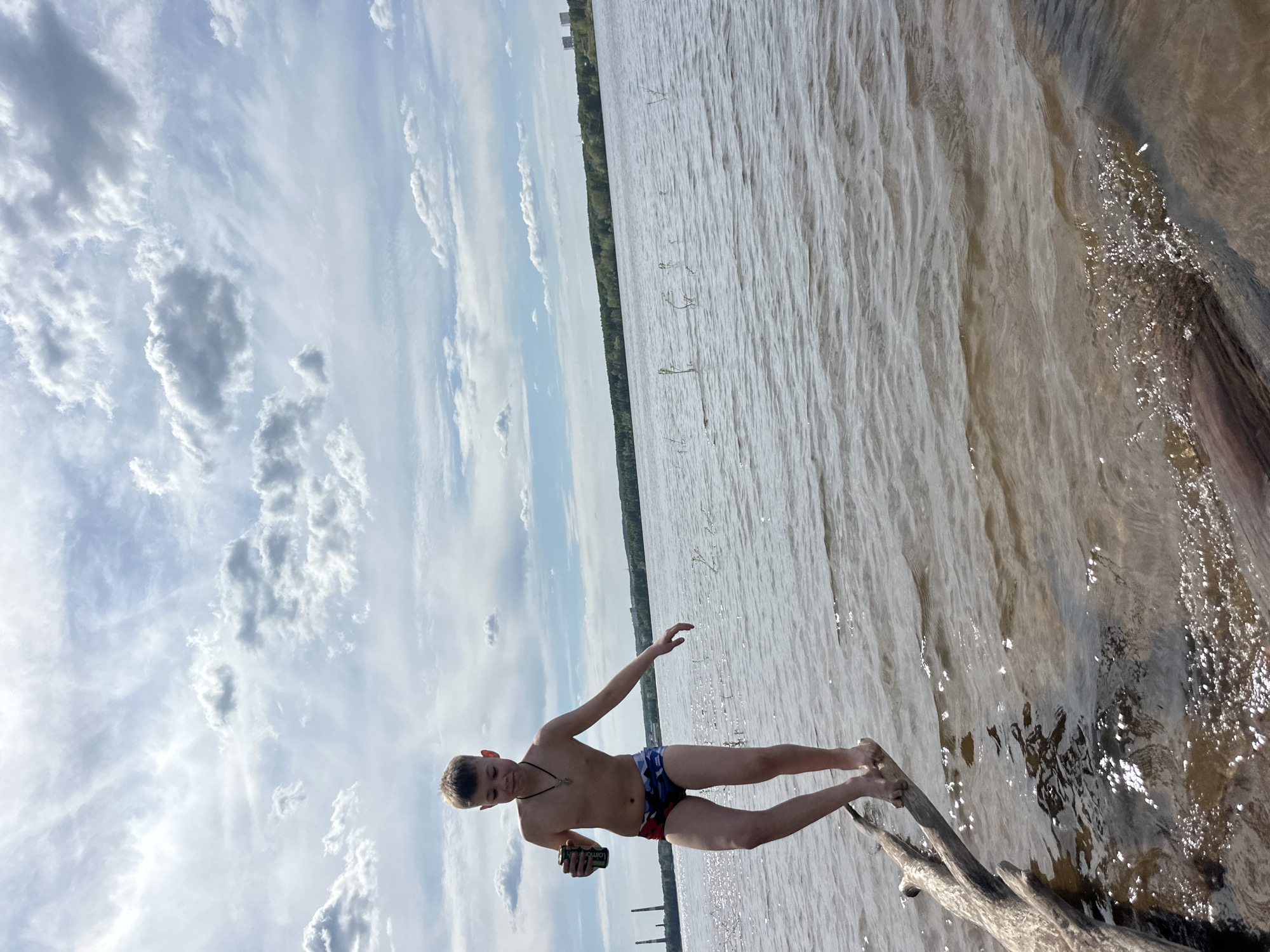 A medium shot captures a person standing on driftwood at the edge of a body of water in the city of Chaikovsky, Russia. The person, possibly male, is fair-skinned, with short brown hair and is wearing swimming trunks. The person is holding a can in their left hand. The body of water is tan and slightly rippled. Beyond the water's edge, a line of trees forms the horizon. The sky is light blue with scattered white clouds.