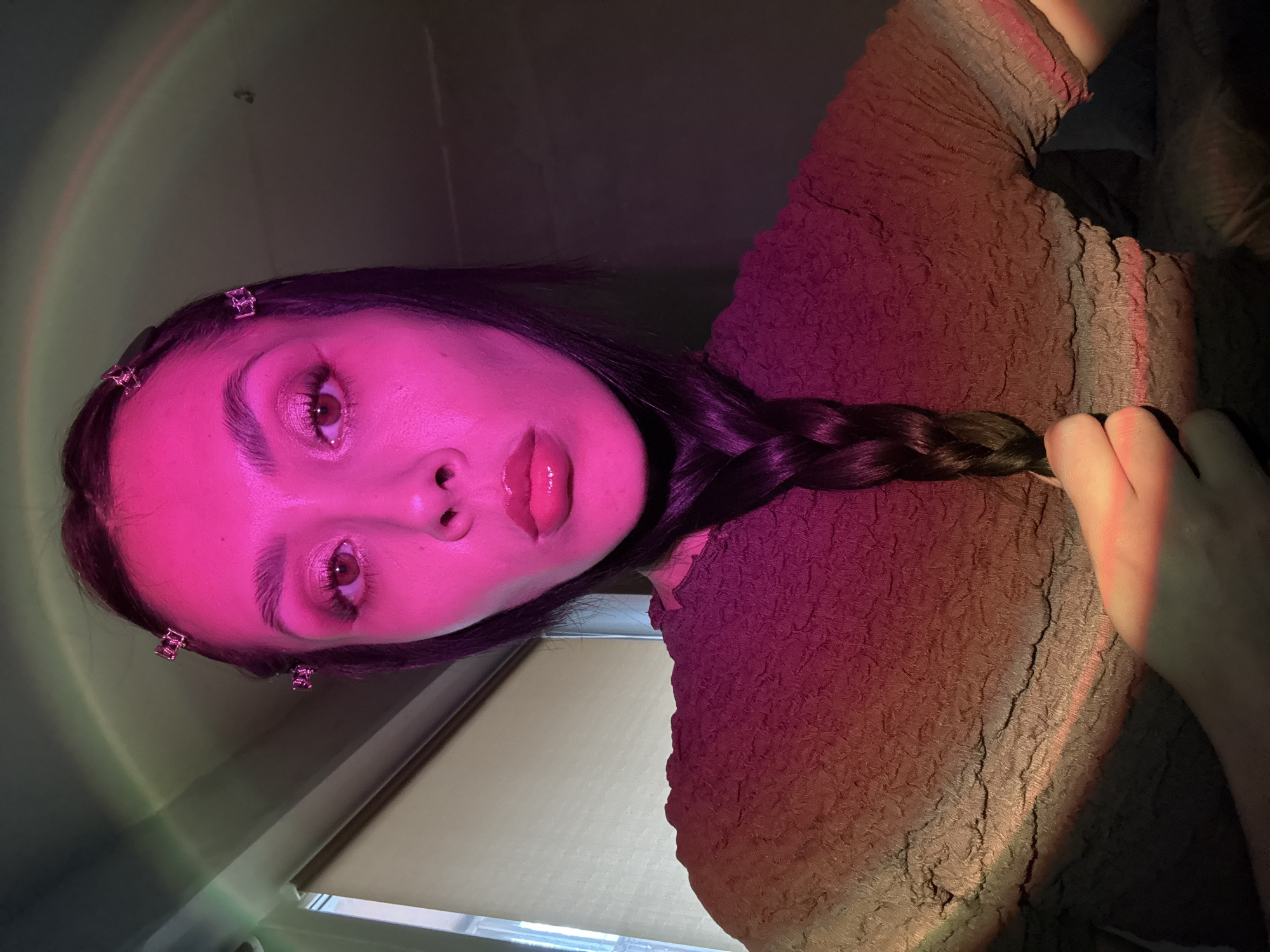 A close-up, tilted portrait of a person with dark hair styled in a braid, facing the camera with a somewhat upward gaze. The person is wearing a brown textured top. There are decorative clips in the hair near the forehead. The image is illuminated by a pinkish-purple light, which casts a rainbow effect on the background. The location is not possible to determine from the given image and the request for Votkinsk, Russia is not supported.