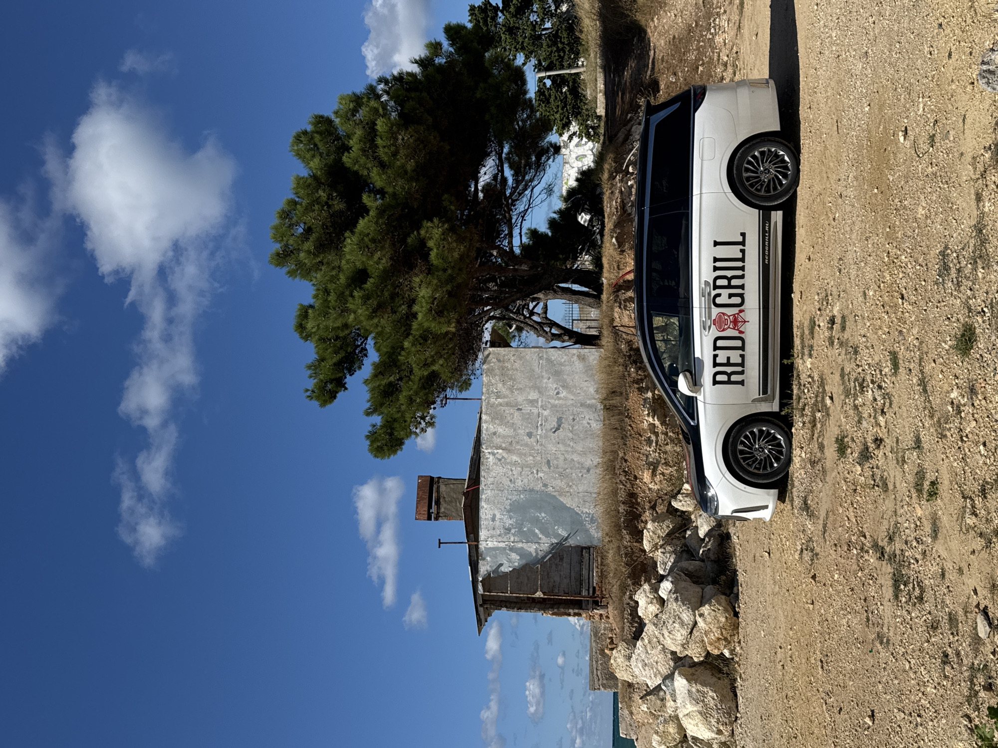 A white van with "RED GRILL" branding is parked on a dirt and gravel lot near the sea. Behind the van is a weathered, dilapidated building with a metal roof and a brick chimney. A large pine tree with dark green needles dominates the middle ground. The sky is blue with scattered white clouds. The scene appears to be located in Sevastopol, Ukraine.