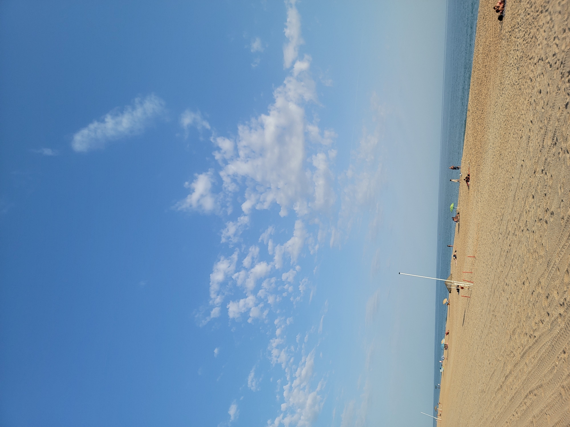 This is a wide-angle, eye-level shot of a beach in Quarteira, Portugal, on a clear, sunny day. The sky is a vibrant blue, dotted with scattered white clouds. The sea, a deep blue, stretches out to the horizon, meeting the sky in a seamless line. The foreground is dominated by a sandy beach, marked with tire tracks and footprints. Several people are visible on the beach, some lounging under umbrellas, others walking or playing. There are also several tall, white poles with flags or markers on them, and a few red posts dotting the sand. The overall atmosphere is peaceful and relaxed, suggesting a typical day at the beach.
