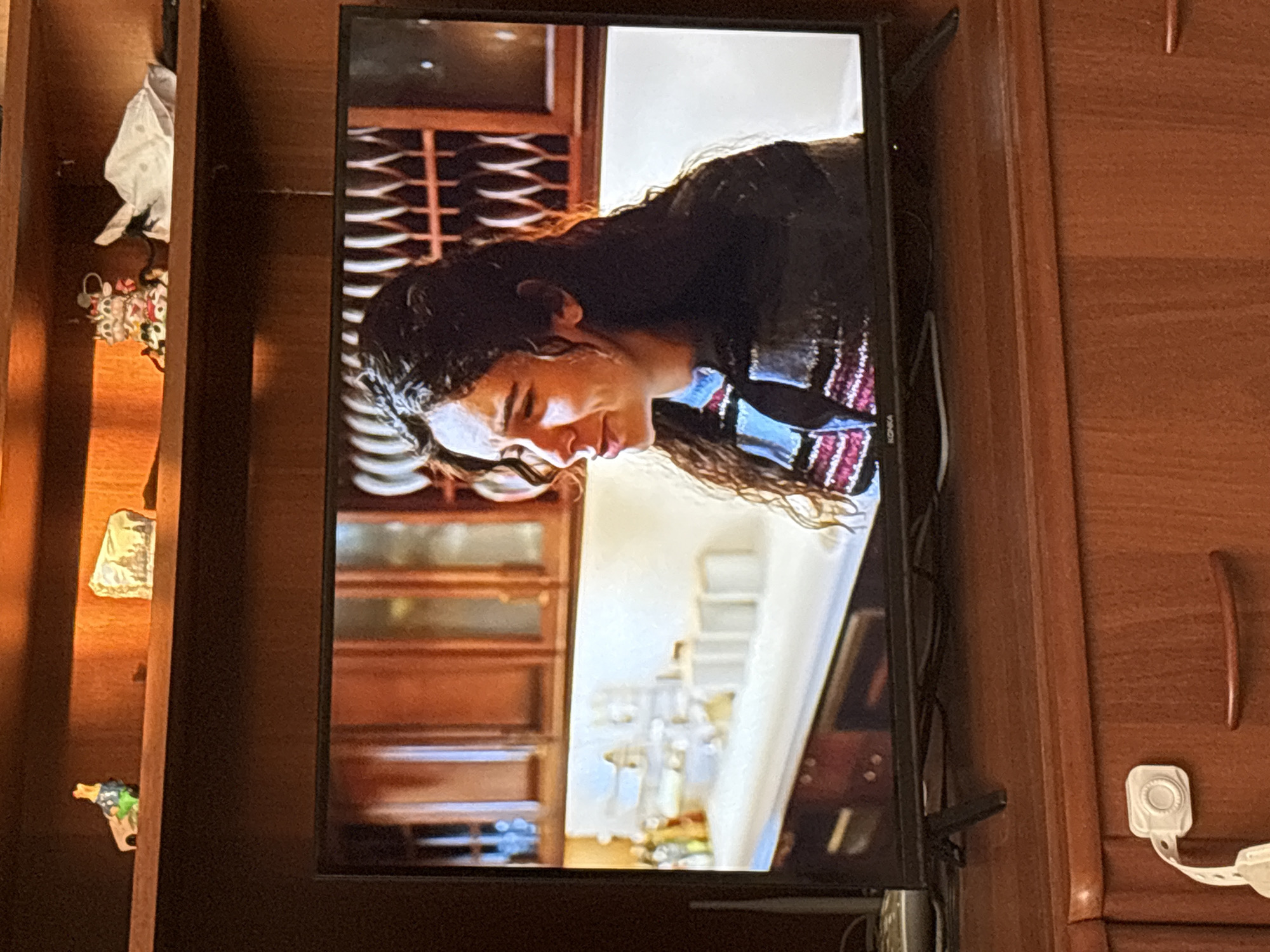 A television screen displays a scene of a young woman with long, dark, curly hair, looking distressed. She is wearing a striped sweater with blue, red, and white stripes. The background on the screen appears to be a kitchen with wooden cabinets and white countertops.
The television is placed on a wooden cabinet. On the shelf above the television, several small decorative items are visible, including a small figurine of a duck or bird, a small rectangular object with a picture of a building, and what appears to be a white folded cloth or piece of paper.
The cabinet itself is made of wood and has a reddish-brown finish. A white childproof lock is attached to the front of one of the drawers. The overall environment suggests a domestic setting. The image is taken from an angle that captures the television screen and the furniture it rests on, with the surrounding shelving.