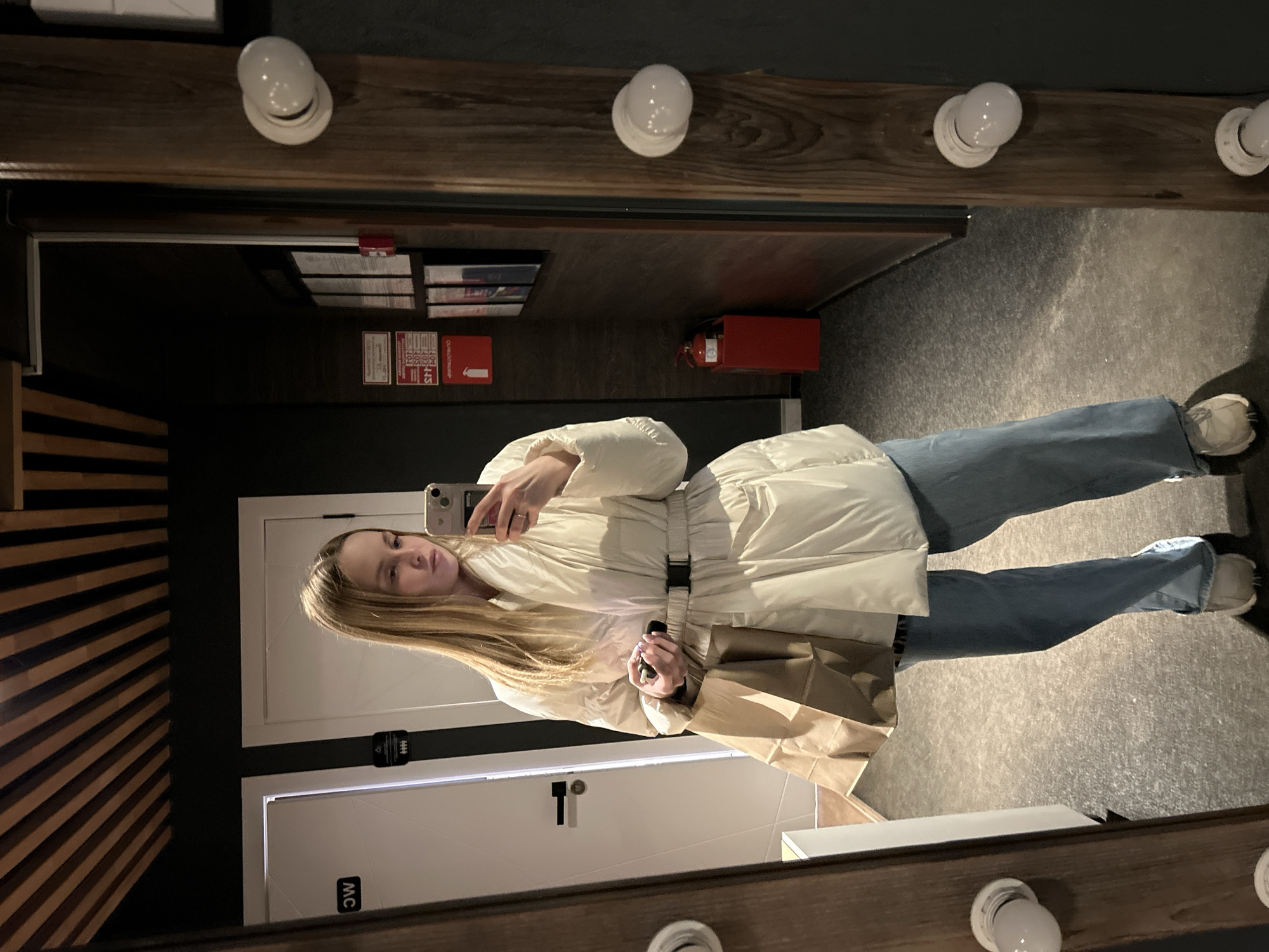 The image is a mirror selfie featuring a young woman standing in an indoor setting. She has long, straight blonde hair and a neutral expression, looking directly at the smartphone she holds in her right hand to take the picture. She is wearing a white or off-white long puffer jacket, belted at the waist with a black buckle, blue jeans, and light-colored sneakers. In her left hand, she holds a brown paper bag. A ring is visible on her right ring finger.
The reflection shows a dark-walled room with a textured, light-colored floor. The mirror itself has a thick wooden frame adorned with multiple round, white light bulbs, typical of a vanity or dressing room. To the left of the woman, a decorative wooden slatted wall is visible in the reflection. To her right, two white doors are present. One door has a black sign with the letters "WC" (appearing upside down in the reflection), indicating a restroom. Another door features a black sign with a symbol of multiple stylized people.
Further back in the reflection, several red signs are mounted on the dark wall. Visible text on these signs includes numbers like "400," "401," "403," "404," and "405," possibly indicating room numbers or floor information. Other red signs display symbols such as an arrow pointing downwards with a person, and a fire symbol, along with partial text "ОГНЕ..." (likely referring to "ОГНЕТУШИТЕЛЬ," meaning fire extinguisher in Russian). A red fire extinguisher box is clearly visible on the wall below these signs. The setting suggests a modern public facility, possibly a hotel corridor, event venue, or commercial building in Chaikovsky, Russia.