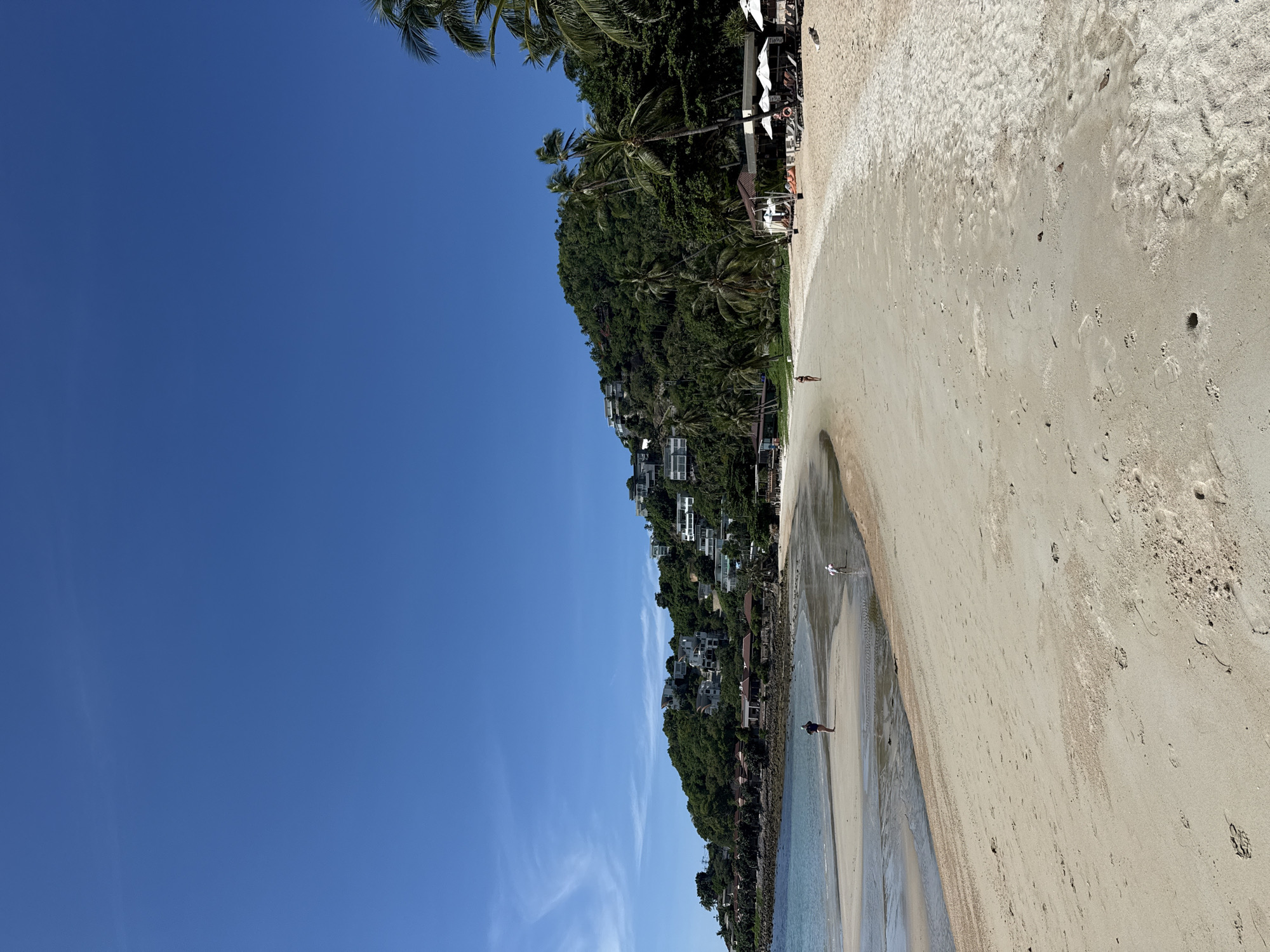 The image depicts a tropical beach scene in Baan Chaweng Noi, Thailand, under a clear blue sky. The foreground is dominated by a wide expanse of sandy beach, marked by footprints and a few small holes. The tide is out, revealing wet sand and shallow pools of water closer to the shoreline.
In the middle ground, two individuals are visible walking on the wet sand. One person, wearing dark shorts and a dark top, stands further out in the shallow water. Another individual in lighter-colored shorts is closer to the waterline. To the right of the beach, several beach umbrellas and lounge chairs are set up, indicating a resort or public area. A small, open-air structure with a thatched roof is visible among the loungers.
The background features a lush, green hillside covered in dense vegetation and palm trees. Several white buildings, appearing to be houses or villas with modern architecture, are nestled into the slope of the hill, suggesting a residential or resort development. The sea, a calm, turquoise color, gently laps against the shore on the left side of the image. Palm trees are scattered throughout the scene, with a few prominent ones framing the right side of the image and extending into the sky. The overall impression is one of a serene and sunny beach day in a tropical paradise.
