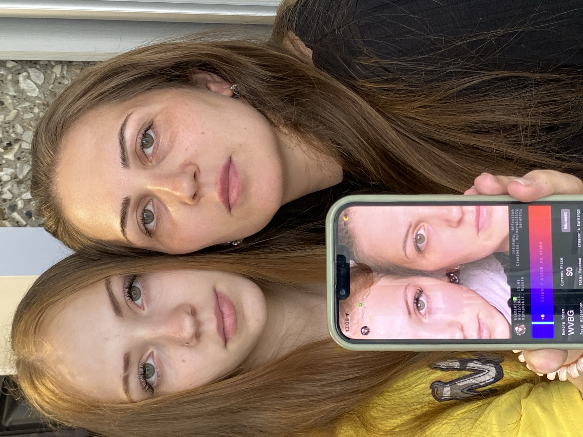 Two young women are closely positioned together, looking at the camera. The woman on top has brown hair, light skin with freckles, and is wearing a black top. The woman below has blonde hair, light skin, and is wearing a yellow top with a "W" logo. A phone is held up in the foreground, displaying a picture of the same two women. The phone screen also shows text, indicating a location in Марковский, Russia, specifically "ESENIAS9, 04/12/2026. 15:01 Markouskii. Russia". The phone screen displays "Intimacy. Affection. Closeness. Vulnerabilitu. Serenity." as well as the words "Swipe/click to stake. Current Price $0. Total Revenue".