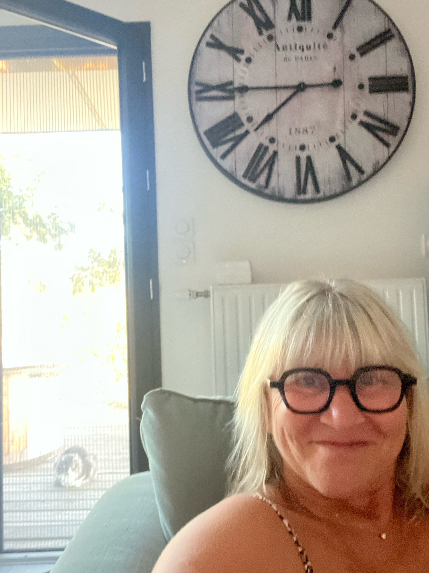 The image is a medium shot selfie taken indoors, looking slightly up at the subject. The subject is a blonde woman with bangs, wearing black-rimmed glasses with a pink tint. She has a light complexion and is smiling. She is wearing a patterned top with a delicate necklace. In the background, a large round clock with Roman numerals and "Antiquité de Paris 1887" is mounted on a white wall. To the left of the subject, a large doorway or window with a dark frame reveals an outdoor scene with bright sunlight and foliage. A light-colored radiator is visible below the clock. The overall impression is a casual indoor setting, possibly a living room or hallway. No specific location within Chambœuf, France is discernible from the image.