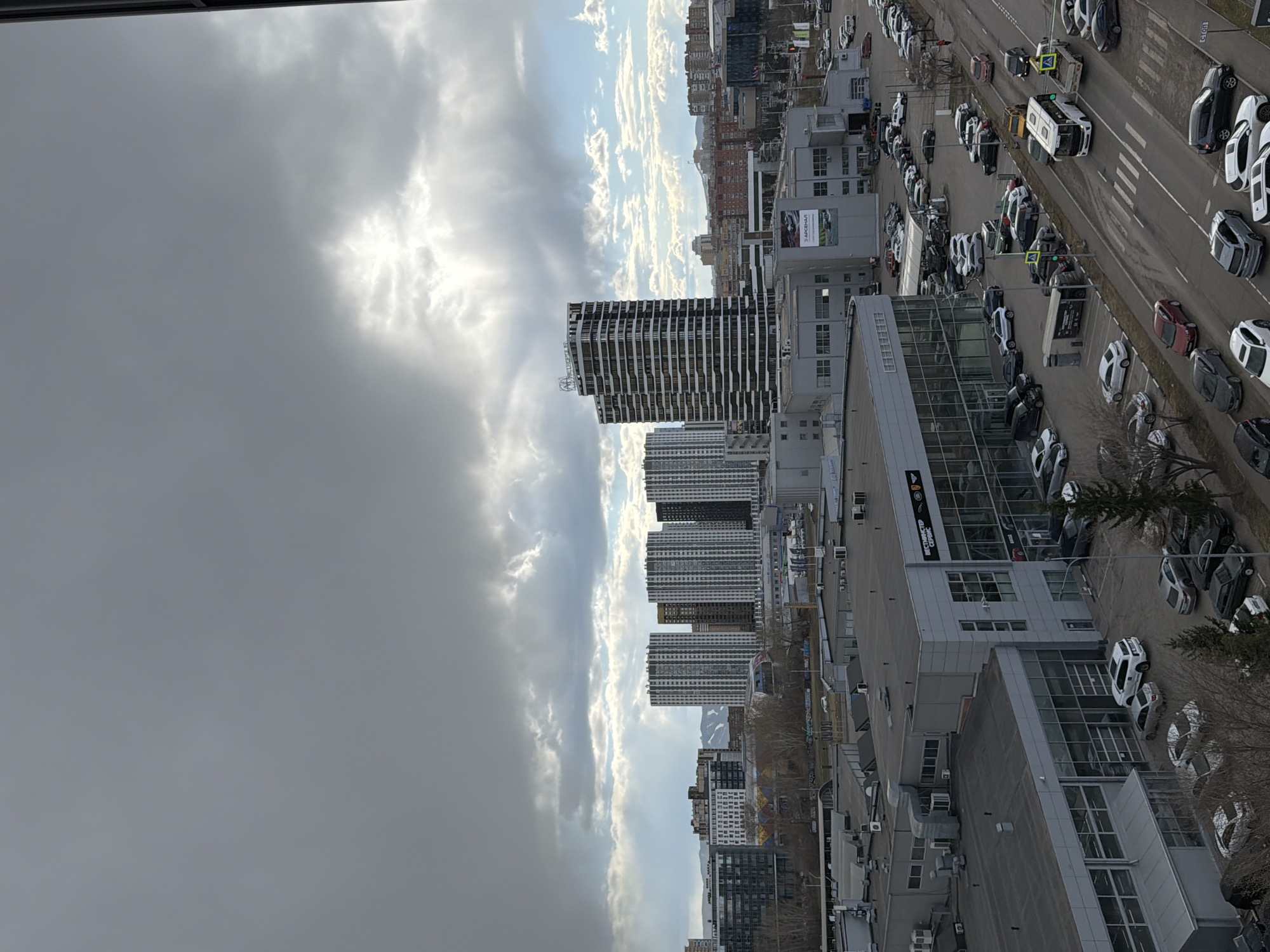 This high-angle, panoramic image captures an urban landscape under a dramatic, cloudy sky, likely in Krasnoyarsk, Russia, given the location context and visible Cyrillic text. The perspective is looking over a sprawling cityscape dominated by buildings and numerous vehicles.
The sky is a prominent feature, with thick, grey clouds covering much of the upper left portion, while patches of bright sunlight break through in the upper right, illuminating distinct white clouds. Further in the distance, a lighter blue sky is visible, suggesting varying weather conditions.
The foreground and midground are filled with a mix of commercial and residential structures. Several modern, tall buildings with distinctive black and white striped facades rise above the surrounding architecture. Below these are numerous lower-rise, grey commercial buildings, some with large glass windows. A multi-lane road runs through the scene, bordered by vast parking lots.
Cars are a dominant element, with a high concentration of vehicles parked in rows on dealership lots and along the roadside. Many appear to be new cars, predominantly white and dark-colored. Traffic is also visible on the road, including trucks and cars. Pedestrian crossings are marked on the asphalt. Sparse, bare trees are scattered throughout the scene, suggesting late winter or early spring.
Visible text on one of the commercial buildings includes "ВЕСЧЕСТЕР СЕРВИС" (VESCHESTER SERVICE) and "LAND ROVER", indicating a Land Rover dealership or service center. Another sign displays "АРСЕНАЛ" (ARSENAL) next to a car image. The scene depicts a bustling commercial and residential area during daytime, with the dynamic sky adding to the atmosphere.